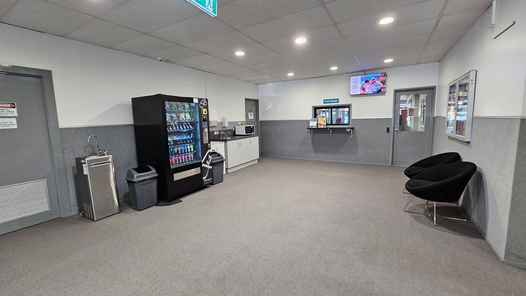 A waiting area with a grey carpeted floor and a tiled ceiling with recessed lights. On the left, there's a water fountain, a vending machine, and a waste bin. A countertop with a microwave and cabinets is nearby. In the centre, a window labelled "Centre Manager" is set into the wall. On the right, two black chairs are positioned against the wall. A television is mounted above the manager's window, and a door is visible on the far right.