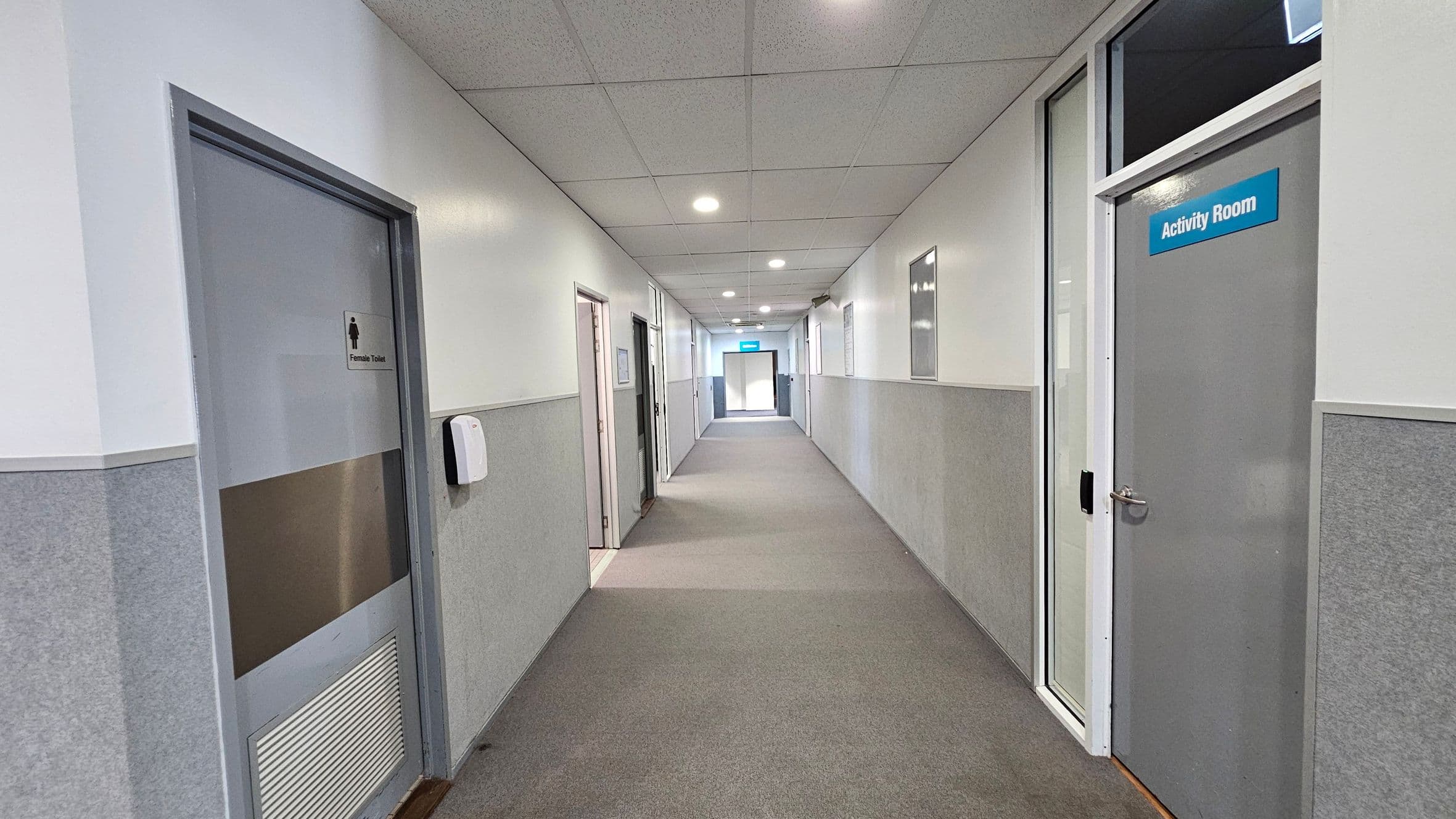 A corridor with grey carpeted flooring and a tiled ceiling with recessed lights. On the left, there is a door labelled "Female Toilet" and a wall-mounted dispenser. On the right, a door marked "Activity Room" is visible. The walls are half-panelled with grey and white. At the end of the corridor, a blue sign is visible above a door, indicating an exit or another room.