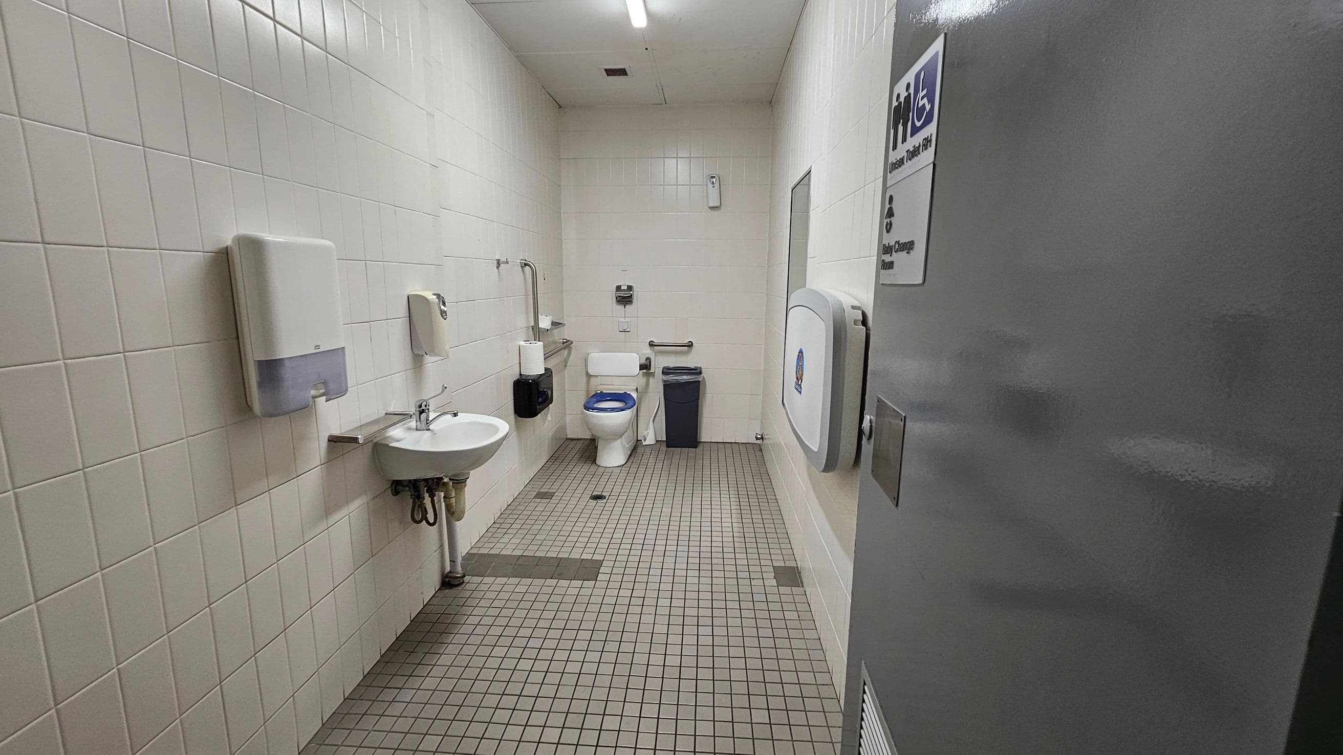 Accessible bathroom with white tiled walls and a green tiled floor. On the left, a sink with a soap dispenser and paper towel holder. In the centre, a toilet with grab bars and a bin nearby. On the right, a baby changing station is mounted on the wall. The ceiling is white with a fluorescent light. A sign on the door indicates it is a unisex and baby change facility.