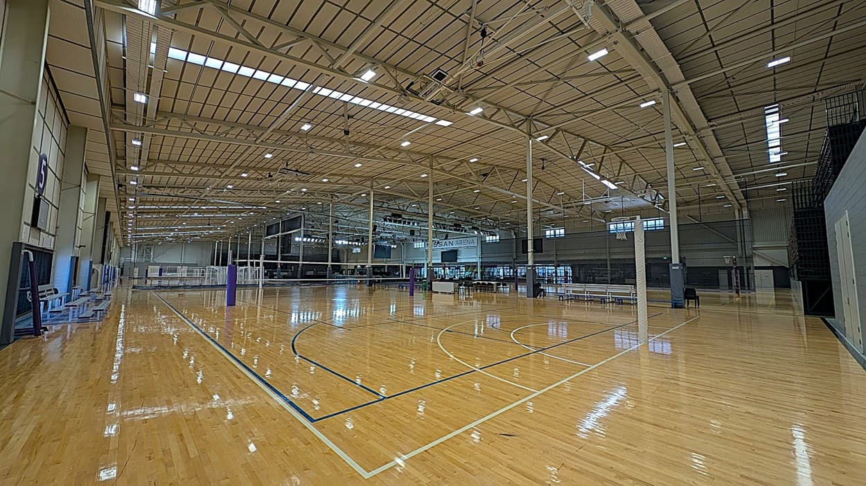 A large indoor sports hall with a polished wooden floor. Multiple netball courts are marked with blue and white lines. Several netball hoops are positioned throughout the space. The ceiling is high with exposed beams and numerous lights. On the left, there is a dark wall with a scoreboard. On the right, benches and equipment are lined up against the wall. In the background, a banner with "NISSAN" is visible above a seating area.