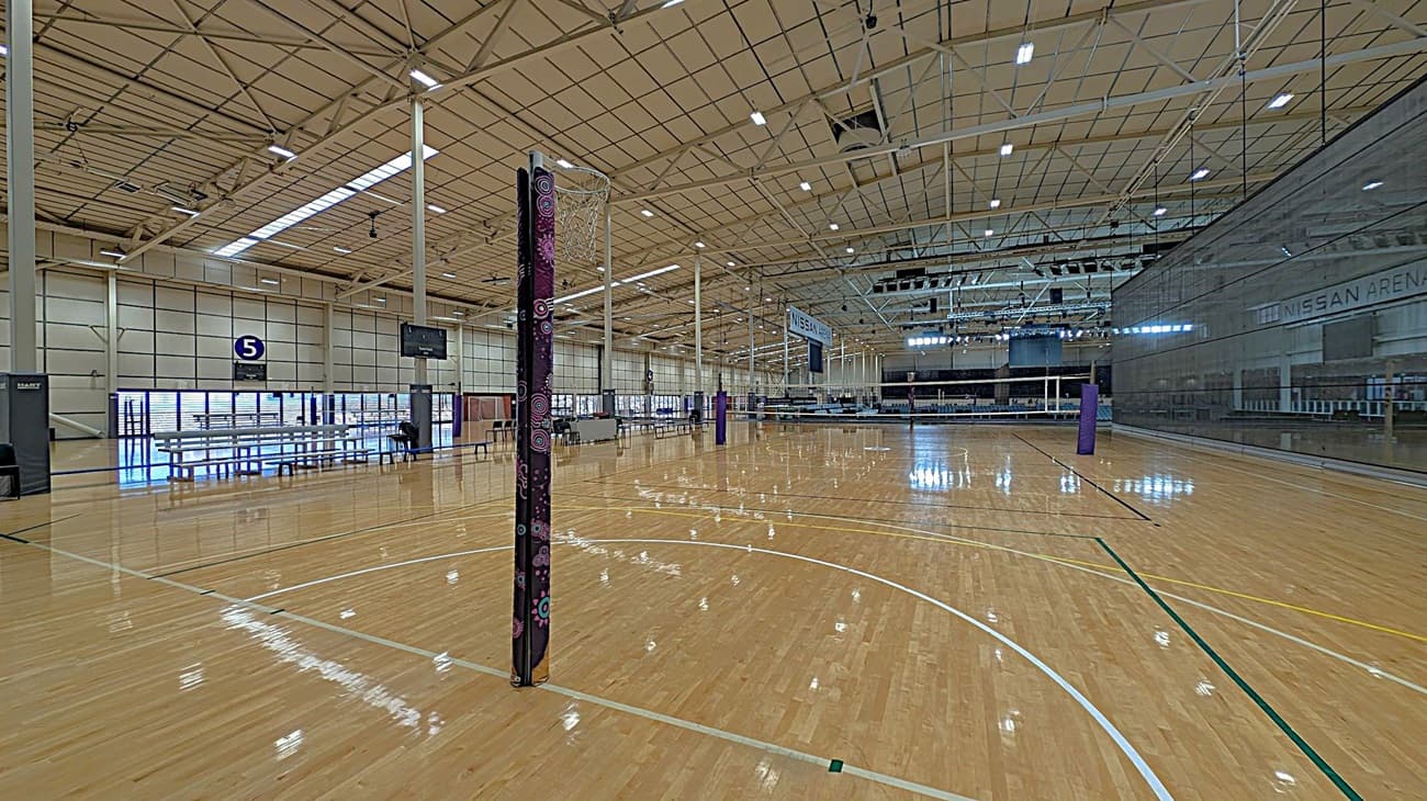 A spacious indoor sports arena with a polished wooden floor marked for netball. In the centre, a netball post with a protective cover stands prominently. On the left, there are rows of benches and a scoreboard. The ceiling is high with exposed beams and lighting fixtures. On the right, a large glass wall with "Nissan Arena" written on it. The background features more seating and additional netball posts.