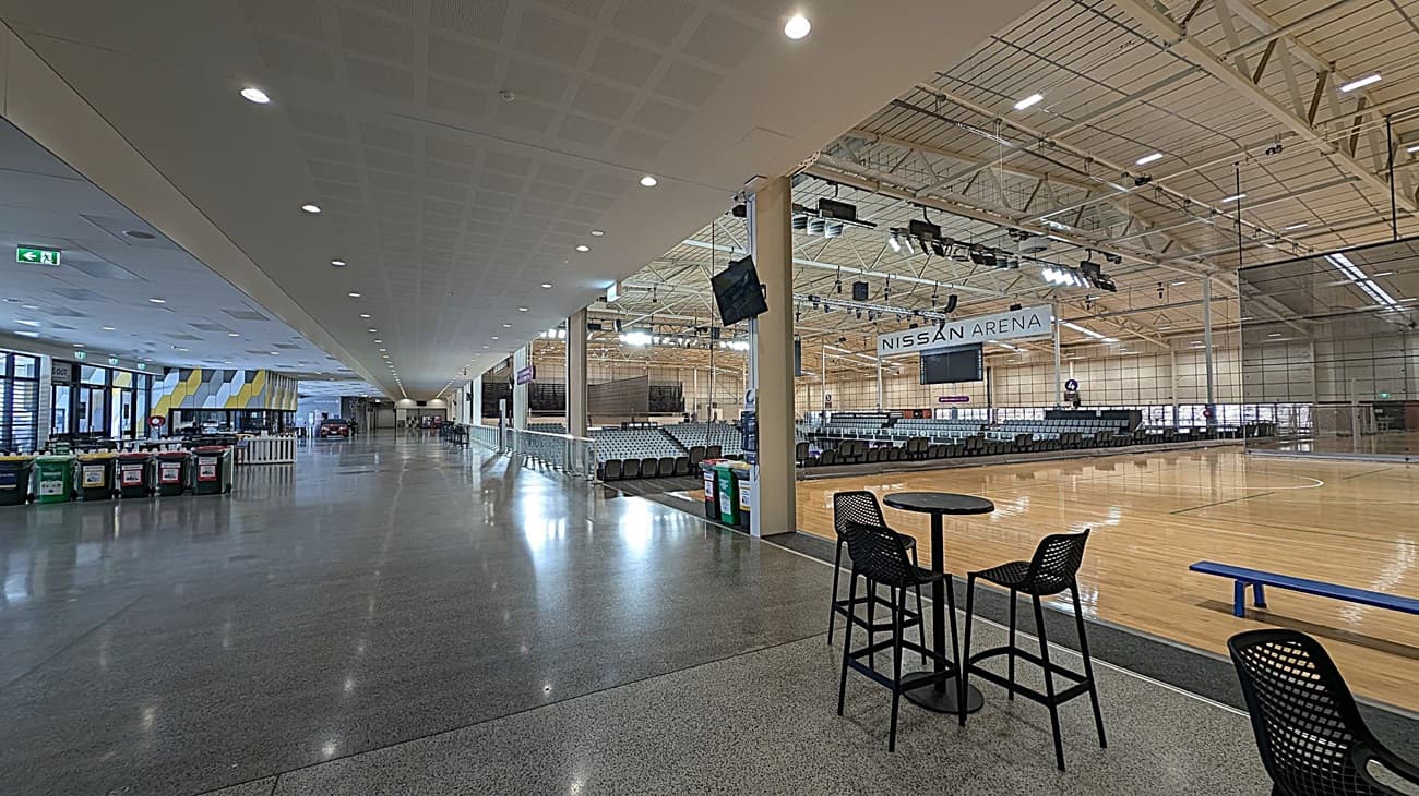 A spacious indoor arena with a polished concrete floor. On the left, there is a wide corridor with waste bins and a patterned wall. The ceiling is lined with recessed lights. On the right, the arena features a wooden sports court with rows of seating and a sign reading "Nissan Arena." A small table with chairs is positioned near the court. The ceiling above the court has exposed beams and lighting fixtures.