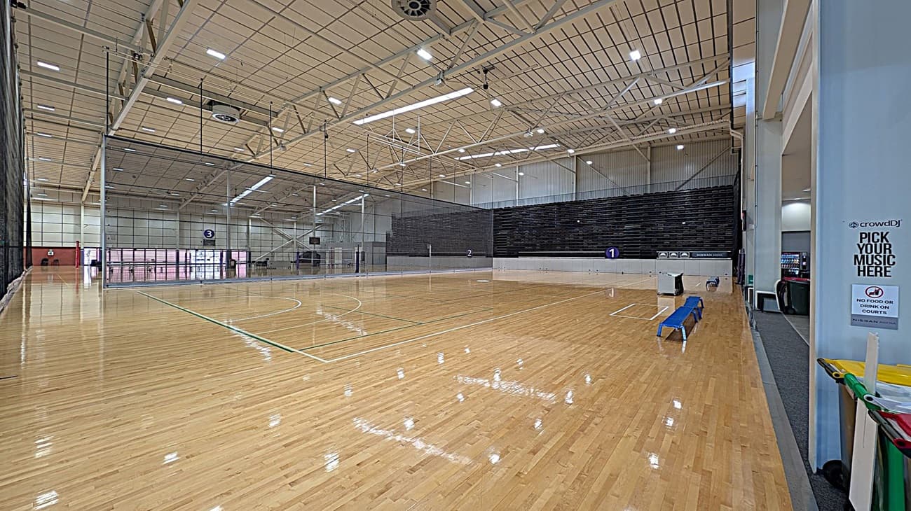 A spacious indoor sports court with a polished wooden floor. On the right, there are blue benches and a sign that reads "PICK YOUR MUSIC HERE" and "NO FOOD OR DRINK ON COURTS." The left side features a large net dividing the court, with a scoreboard visible in the background. The ceiling is high with exposed beams and bright lighting. Vending machines are visible in the far right corner.