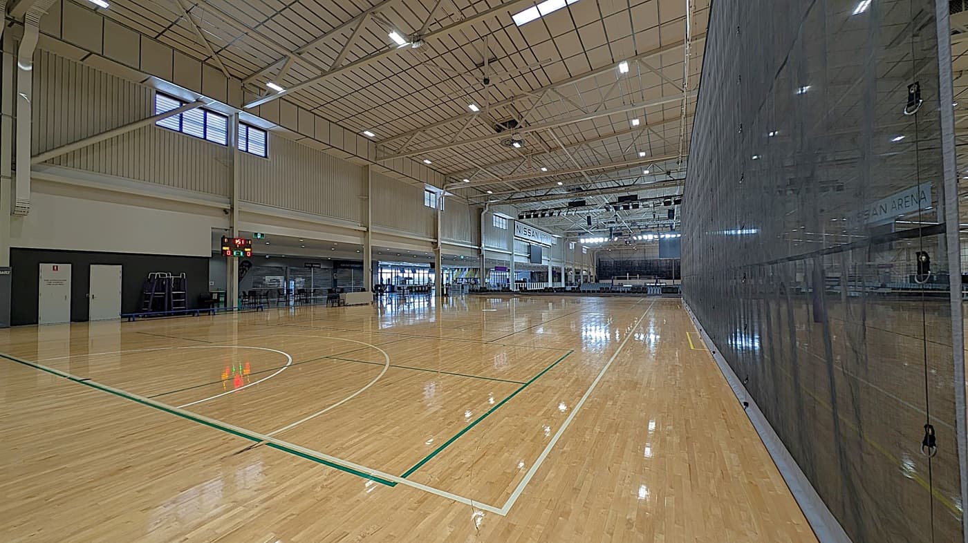 A large indoor sports arena with a polished wooden floor marked for basketball. On the left, there are doors and a scoreboard displaying "45:11" and "0-0". The right side features a tall, dark mesh divider. In the background, there are seating areas and a sign reading "NISSAN ARENA". The ceiling is high with exposed beams and bright lights.