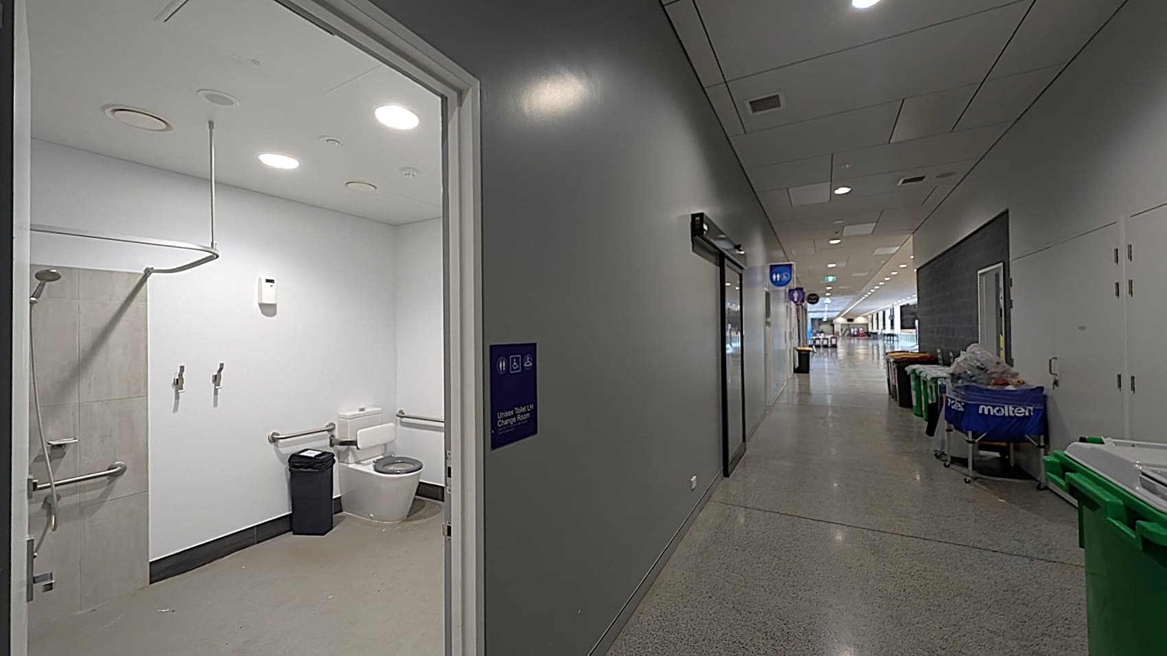A corridor with a polished floor and a ceiling with recessed lighting. On the left, an open door reveals a unisex toilet and change room with a shower, toilet, and handrails. The corridor extends into the distance with several bins and cleaning equipment on the right. Signs indicating facilities are visible along the hallway.