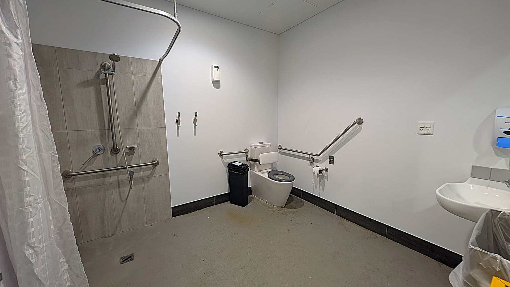 A bathroom with accessibility features. On the left, there is a shower area with a curtain and grab bars. In the centre, a toilet with grab bars on both sides and a black bin nearby. On the right, a sink with a soap dispenser above it. The floor is grey, and the walls are white. The ceiling is plain with a light fixture.