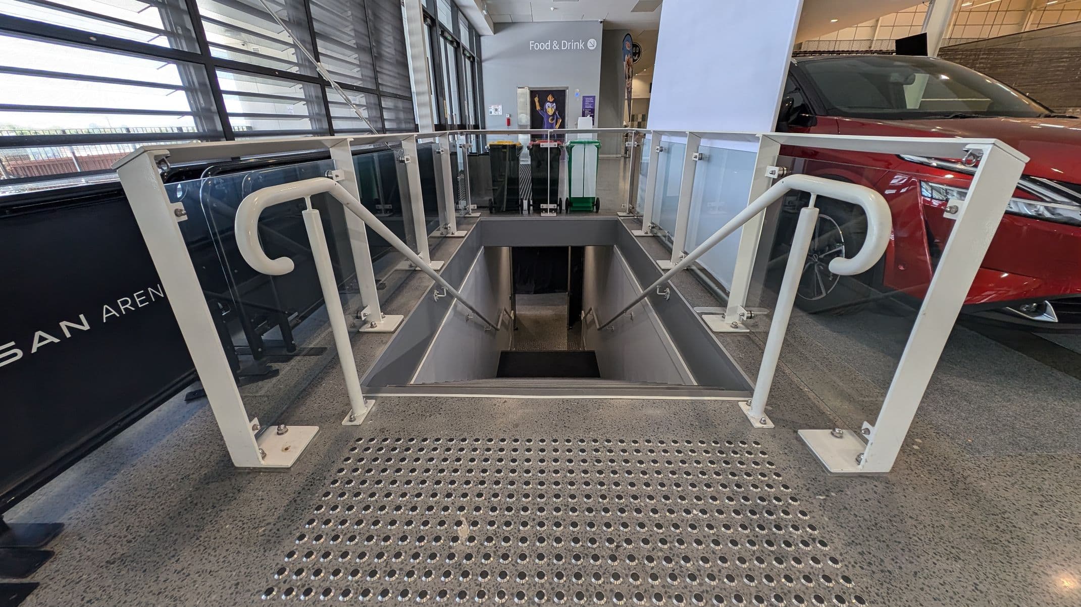 A staircase descends into a lower level, bordered by glass and metal railings. On the left, a black sign with partial text is visible. On the right, a red car is parked. In the background, a sign reads "Food & Drink" with bins and a poster nearby. The floor is speckled with a textured metal mat at the top of the stairs.