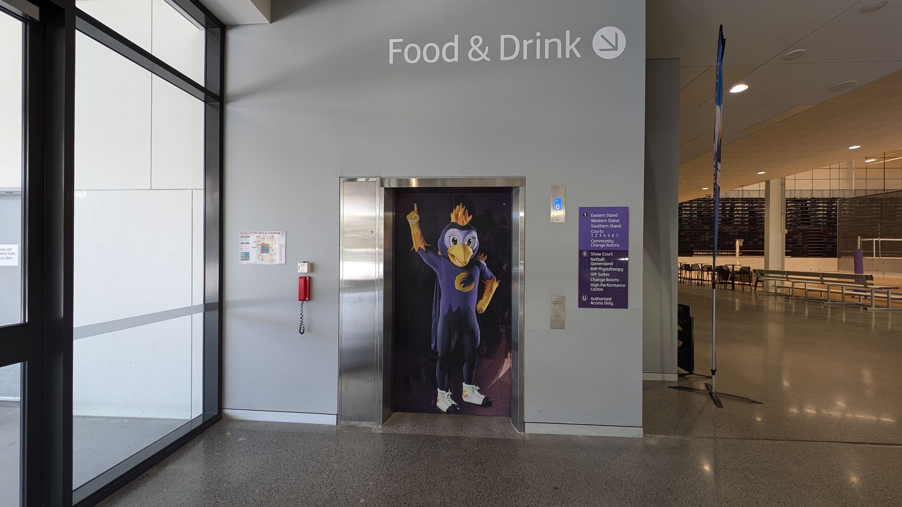A hallway with a lift in the centre, featuring a cartoon bird character on the door. Above, a sign reads "Food & Drink" with an arrow. To the left, large glass windows and a fire extinguisher mounted on the wall. To the right, a sign with directions and a glimpse into a seating area with benches and tables. The floor is polished and reflective, and the ceiling has recessed lighting.