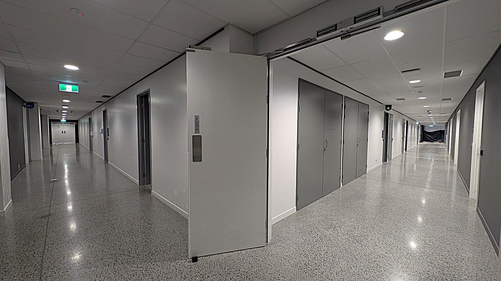 A long, empty corridor with polished, speckled flooring and a white ceiling with recessed lighting. On the left, there are several closed doors and an emergency exit sign. In the middle, a partially open door with a "PUSH" sign is visible. On the right, more closed doors line the wall, leading to a distant end where a dark curtain or barrier is present. The corridor is well-lit and has a clean, modern appearance.