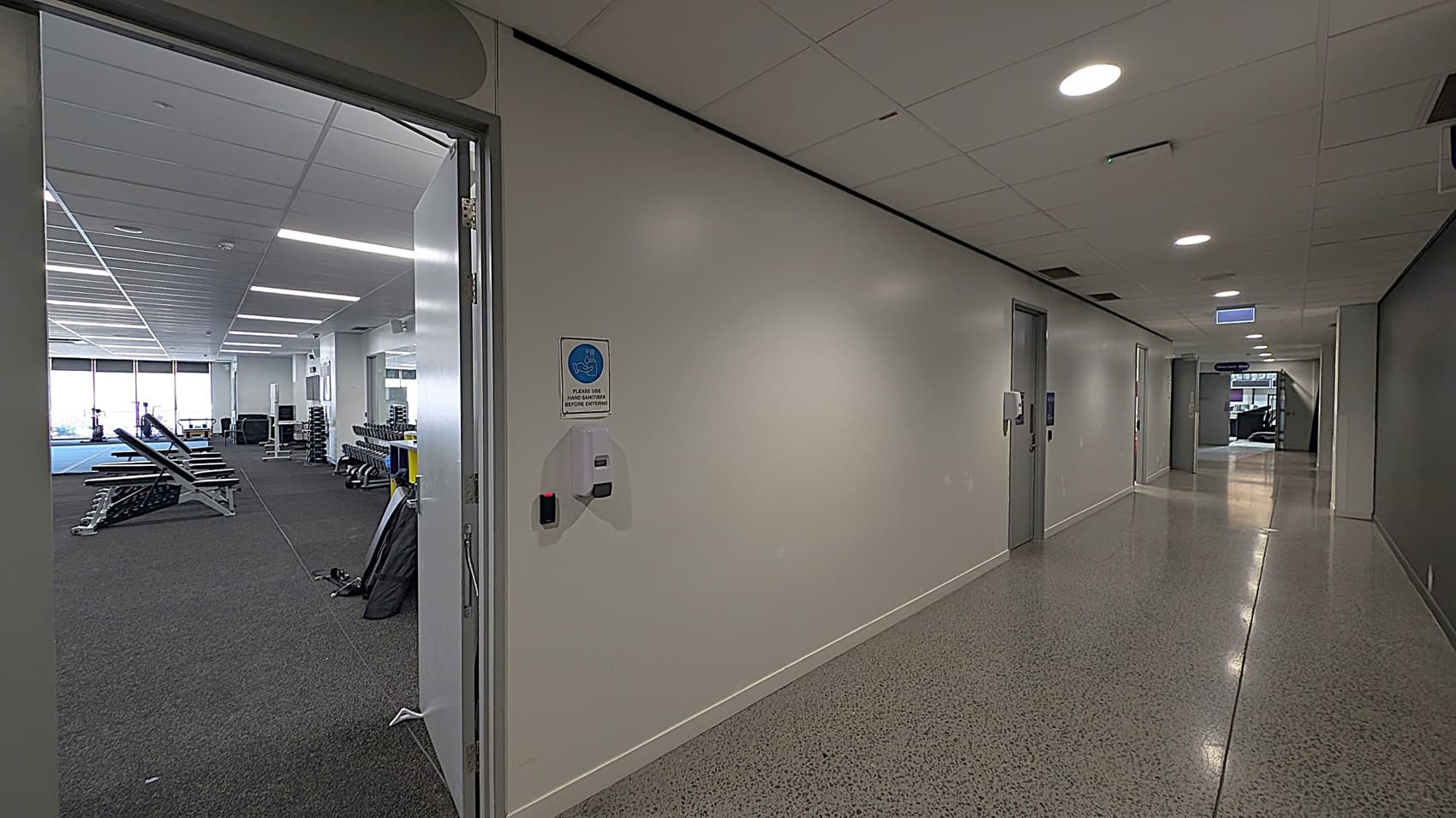 A hallway with a polished, speckled floor and a white ceiling with recessed lighting. On the left, an open door leads to a gym area with exercise benches and weight racks. A sign and hand sanitiser dispenser are mounted on the wall near the door. The corridor extends into the distance with several closed doors on the right and a window at the far end.