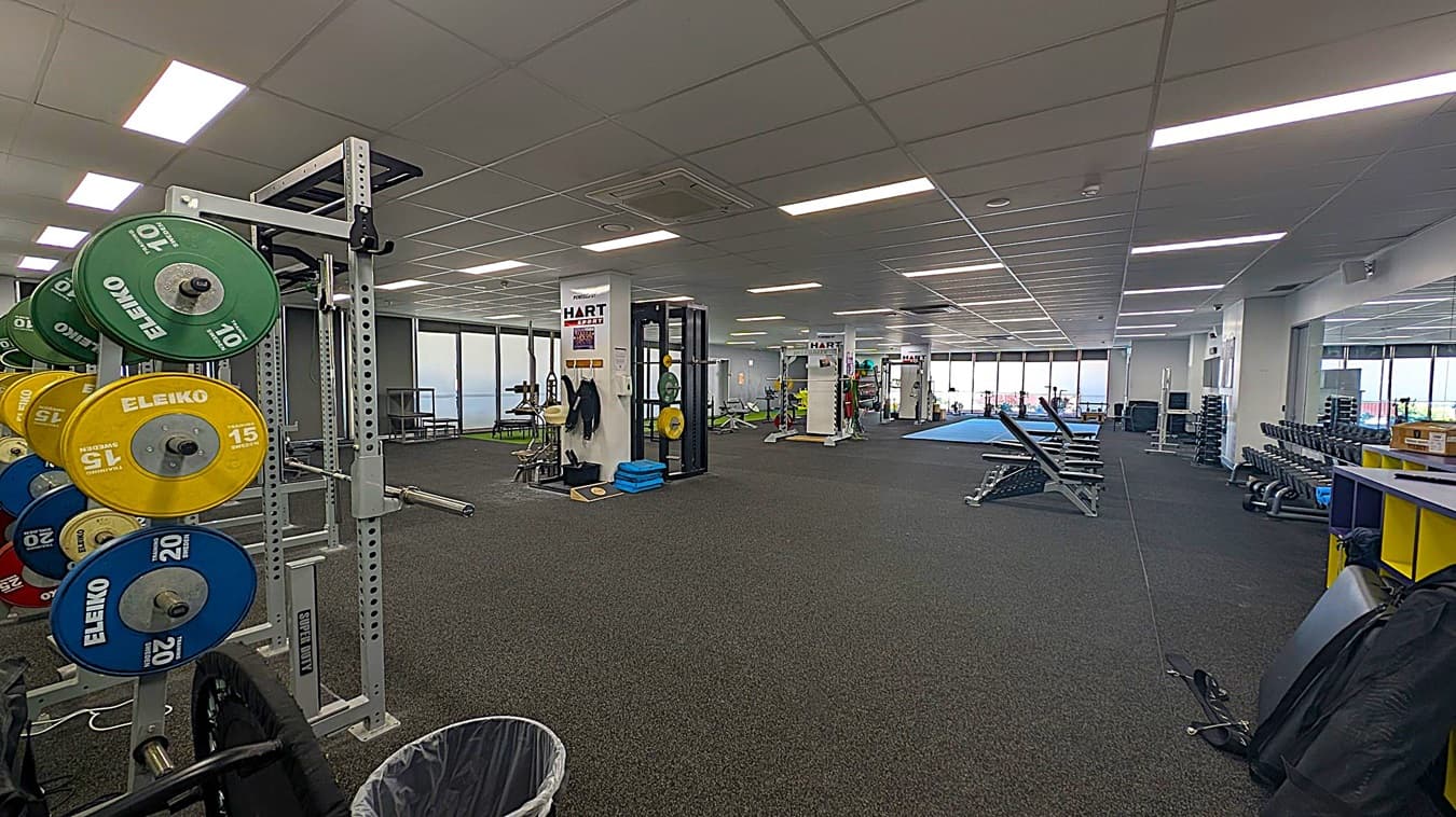 A spacious gym with a variety of equipment. On the left, there are weight plates of different colours and sizes on a rack. In the centre, there are exercise machines and a few benches. The right side has a row of dumbbells on racks and a yellow and blue storage unit. The floor is covered with dark carpet, and the ceiling has a grid pattern with fluorescent lights. Large windows in the background let in natural light.