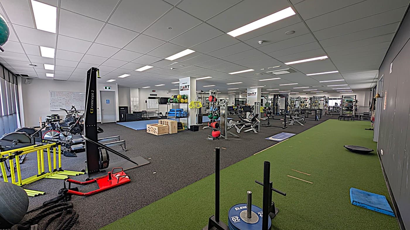 A spacious gym with various exercise equipment. On the left, there are stationary bikes, yellow hurdles, and a rowing machine. In the centre, there are wooden boxes, medicine balls, and weightlifting benches. The right side features a green artificial turf area with weights and a blue mat. The ceiling has bright fluorescent lights, and the floor is a mix of black rubber and green turf. Walls have motivational posters and a whiteboard.