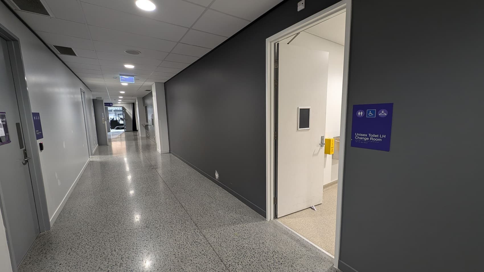 A corridor with a polished, speckled floor and a white ceiling with recessed lights. On the right, a door is open, leading to a unisex toilet and change room, indicated by a blue sign. The walls are painted grey. On the left, there are closed doors and a sign hanging from the ceiling. At the end of the corridor, there is a glass entrance or exit with natural light coming through.