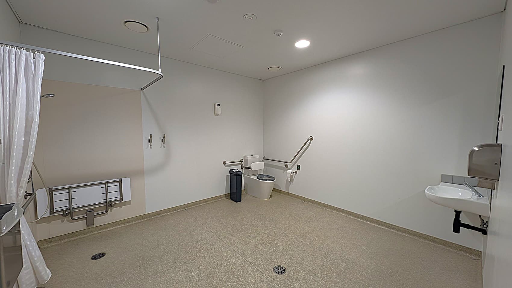 A spacious, accessible bathroom with a green speckled floor and white walls. On the right, there's a wall-mounted sink with a soap dispenser above it. In the centre, a toilet with support rails on both sides and a bin nearby. On the left, a folded wall-mounted changing table and a shower curtain rail. The ceiling is white with recessed lighting and ventilation.