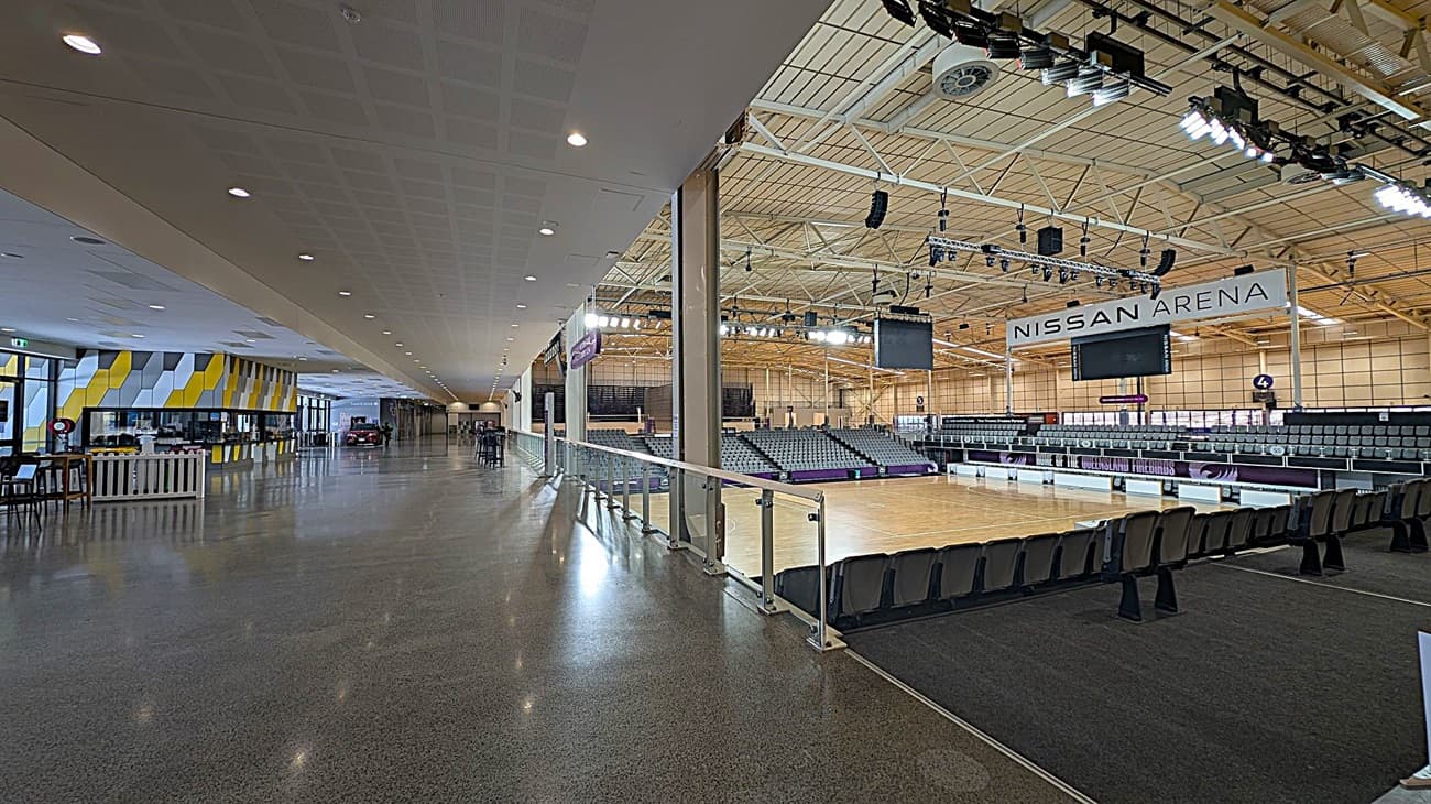 On the left, a spacious corridor with a polished grey floor and a ceiling with recessed lights. The wall features a geometric pattern in yellow, grey, and white. In the middle, a glass railing separates the corridor from the arena. On the right, the arena has tiered seating and a large sign reading "Nissan Arena." The ceiling is high with exposed beams and lighting fixtures.