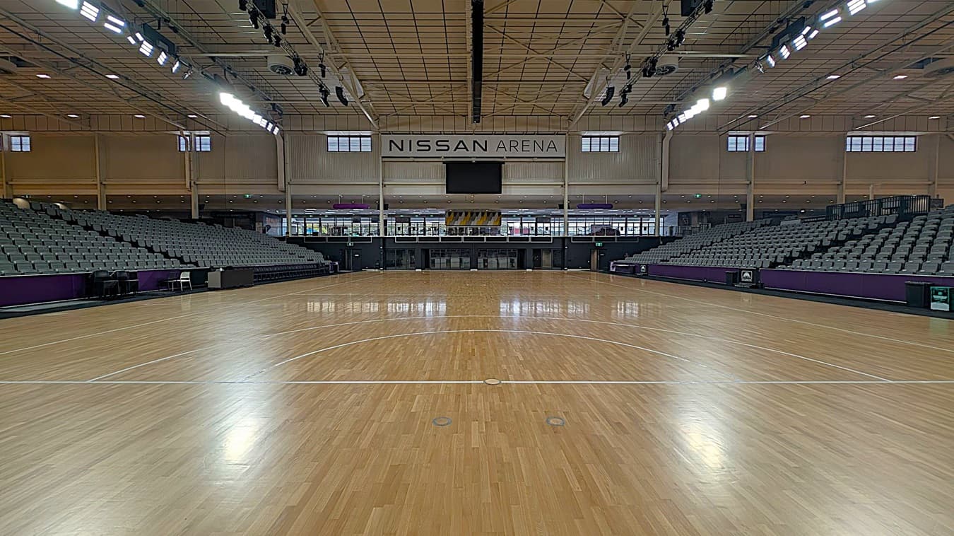 Interior of a large sports arena with a polished wooden floor. On the left and right are rows of grey seating, slightly elevated. The ceiling is high with visible lighting fixtures. At the far end, a sign reads "NISSAN ARENA" above a central entrance. The walls have windows near the ceiling, allowing natural light. The arena is empty, with no people present.