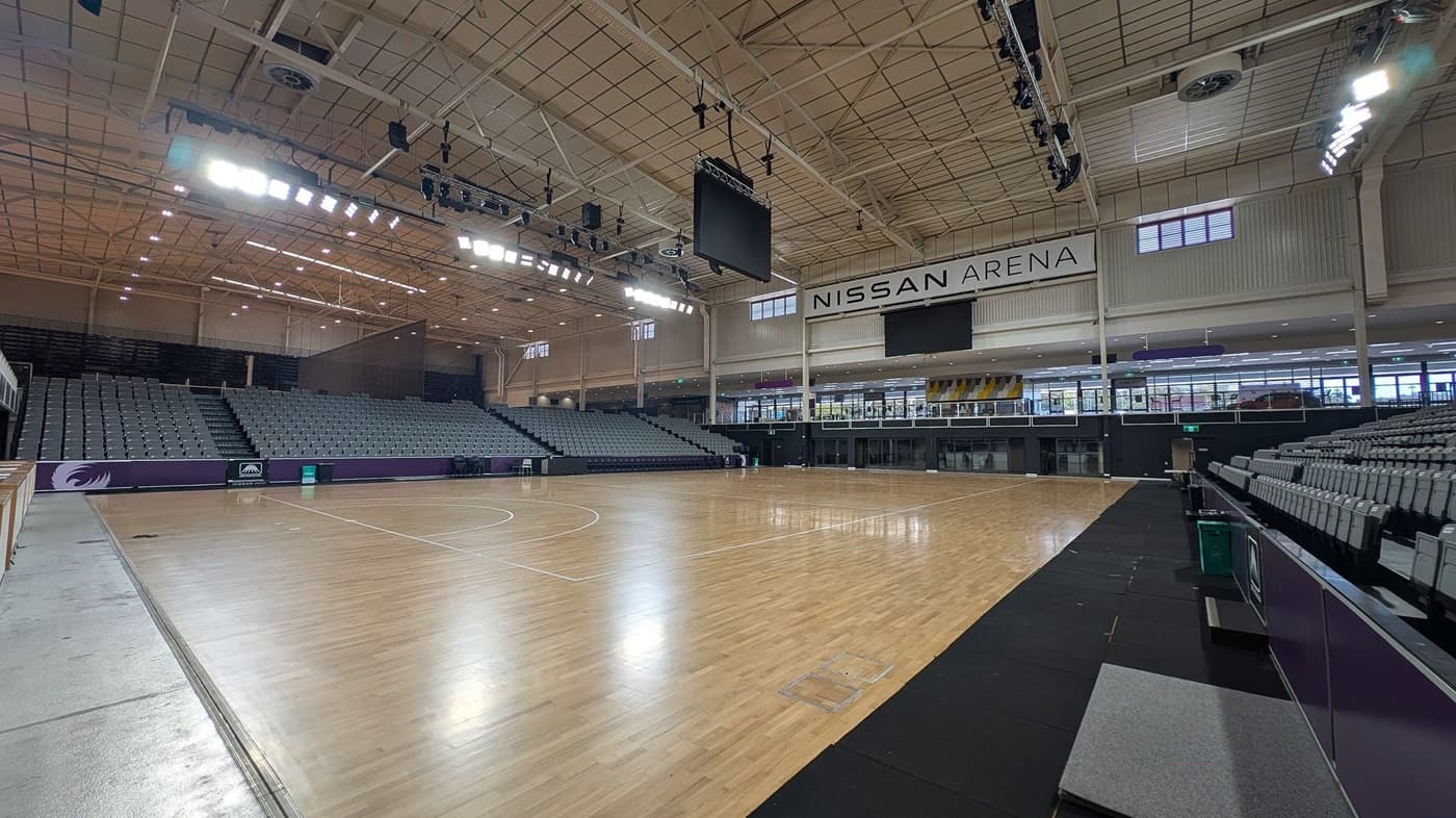 A large indoor sports arena with a polished wooden floor. On the left, there are tiered seating areas with grey seats. The right side also features similar seating. In the centre, a large scoreboard hangs from the ceiling. The ceiling is high with exposed beams and lighting fixtures. The back wall displays the name "Nissan Arena" above a row of windows and doors. The arena is well-lit, and the space is open and spacious.
