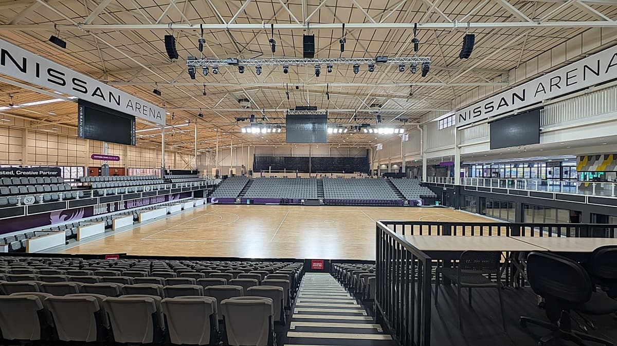 Interior of a large sports arena with tiered seating on both sides and a wooden court in the centre. The ceiling is high with exposed beams and lighting fixtures. Large screens and banners with "Nissan Arena" are visible. The foreground shows a railing and a few chairs. The seating is grey, and the arena is well-lit with natural and artificial light.