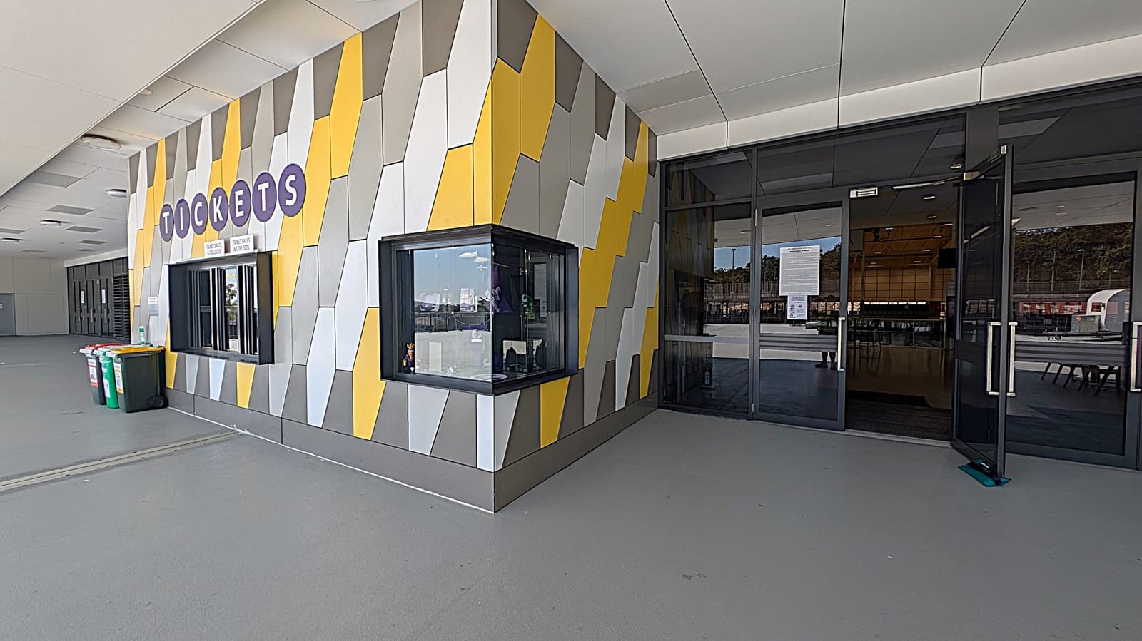 A ticket booth with a colourful geometric wall pattern in shades of yellow, grey, and white. The word "TICKETS" is displayed in large letters above the window. To the right, there are glass doors with handles, leading into a building. The floor is grey and smooth, and there are several bins to the left of the booth. The ceiling is white with recessed lighting.