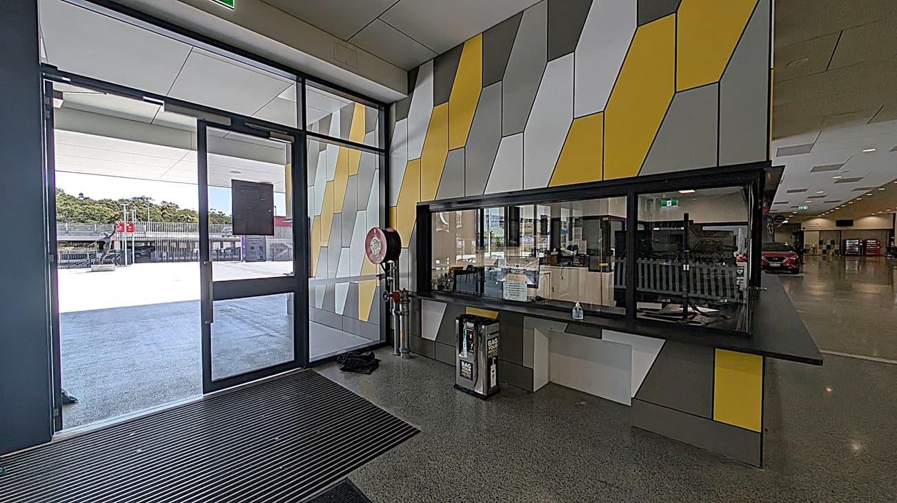 A modern interior with a geometric-patterned wall in shades of grey and yellow on the right. A large glass door on the left opens to an outdoor area with a view of trees and a building. Inside, there's a counter with a glass partition, a fire hose reel, and a bag disposal unit. The floor is speckled grey, and the ceiling has recessed lighting. In the background, a hallway leads to a space with parked cars and vending machines.