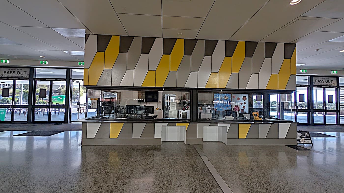 A modern interior with a geometric-patterned counter and wall in shades of yellow, grey, and white. The counter has a service window with equipment inside. The floor is polished concrete with green accents. In the background, there are large glass doors with "PASS OUT" signs above them, leading to an outdoor area with visible seating and greenery. The ceiling is composed of large, light-coloured panels with recessed lighting.