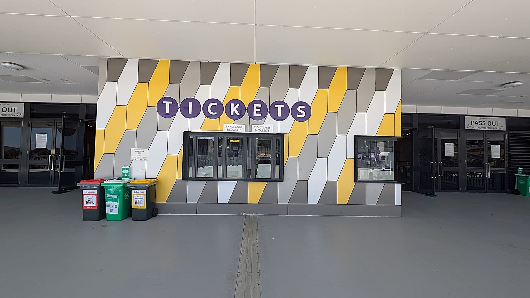 A ticket booth with a colourful geometric wall pattern featuring yellow, grey, and white shapes. The word "TICKETS" is displayed in large blue circles above the window. To the left, there are three bins with red, green, and yellow lids. On either side of the booth, there are glass doors with signs above them, one reading "PASS OUT". The floor is grey, and the ceiling is light-coloured with circular fixtures.