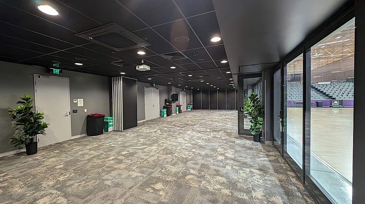 A corridor with a patterned carpet in shades of green and beige. On the left, there are grey doors and potted plants. Several bins are placed along the wall. The ceiling is black with recessed lighting. On the right, large glass windows offer a view into an adjacent area with tiered seating.