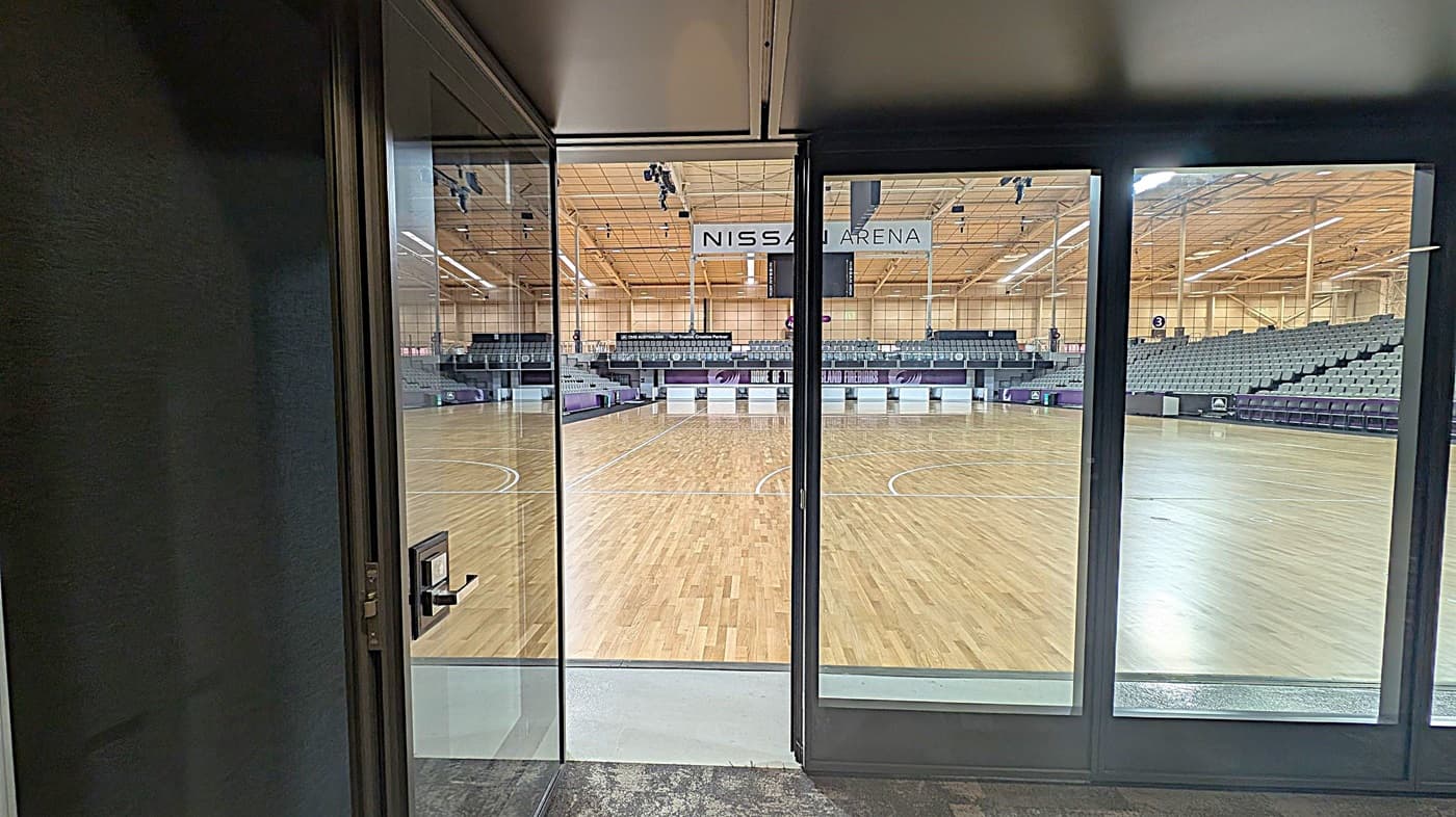 View through glass doors into an indoor sports arena with a wooden floor. The arena has tiered seating on both sides, with a large sign reading "NISSAN ARENA" hanging from the ceiling. The ceiling is high with visible lighting fixtures. The floor in the foreground is dark and textured, contrasting with the bright arena interior.