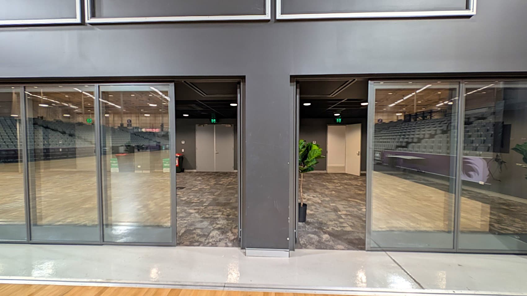 Glass doors with metal frames are in the centre, leading to a room with patterned carpet and a potted plant. On the left and right, there are reflections of an indoor sports arena with tiered seating. The ceiling has recessed lighting, and the floor in the foreground is shiny and smooth.
