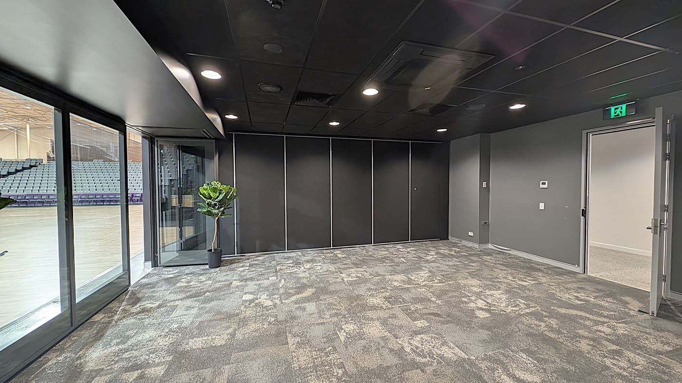 A room with a patterned carpet in shades of grey and green. On the left, large glass windows reveal a view of an adjacent area with rows of blue seats. A potted plant is near the windows. The back wall is dark with panelled sections. On the right, a door is open, leading to another room. The ceiling is dark with recessed lighting, and an illuminated exit sign is above the door.
