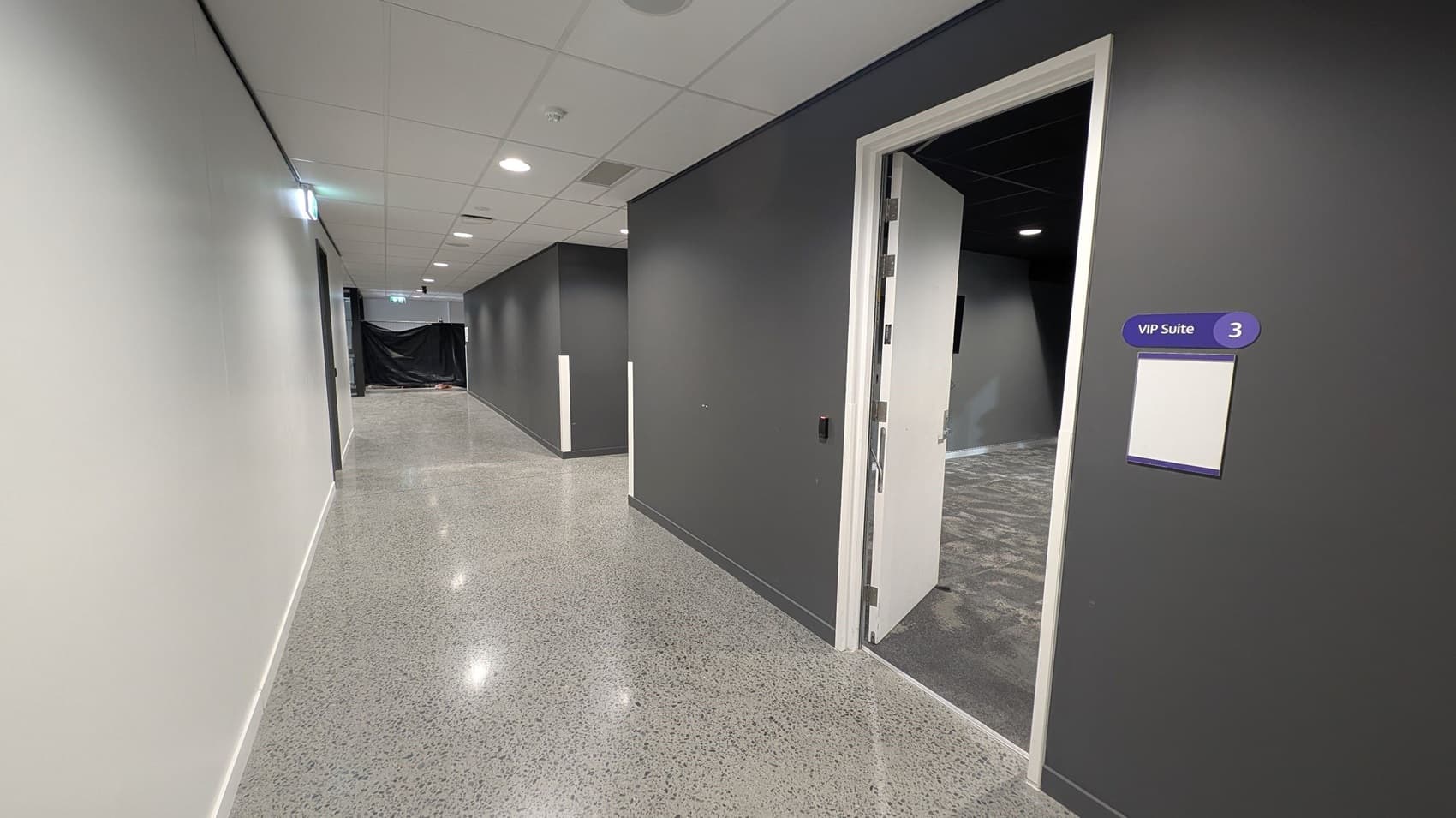 A corridor with polished speckled flooring and a white ceiling with recessed lights. On the right, a door is open, leading to a room labelled "VIP Suite 3" with a sign on the wall. The walls are painted grey. In the background, the corridor extends with a black curtain partially visible at the end.