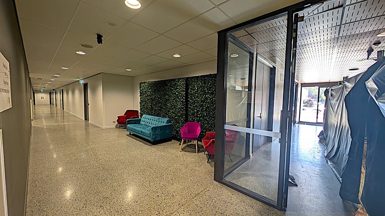 A hallway with a polished, speckled floor and a ceiling with recessed lighting. On the left, a long corridor extends with several doors. In the middle, a seating area features a blue sofa and two red chairs against a wall with green foliage. On the right, a glass door leads to another area with covered items and an exit visible in the background.