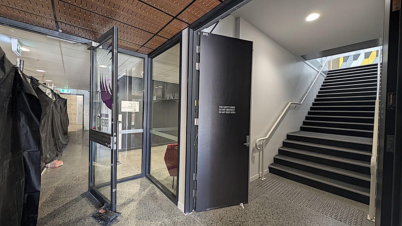 A hallway with a glass door on the left, partially open, leading to a corridor with black draped coverings. A sign on the door reads "FIRE SAFETY DOOR DO NOT OBSTRUCT DO NOT KEEP OPEN." On the right, a staircase with a white handrail ascends, with a patterned wall visible at the top. The ceiling is wooden with a grid pattern, and the floor is speckled. A round ceiling light illuminates the stairway area.