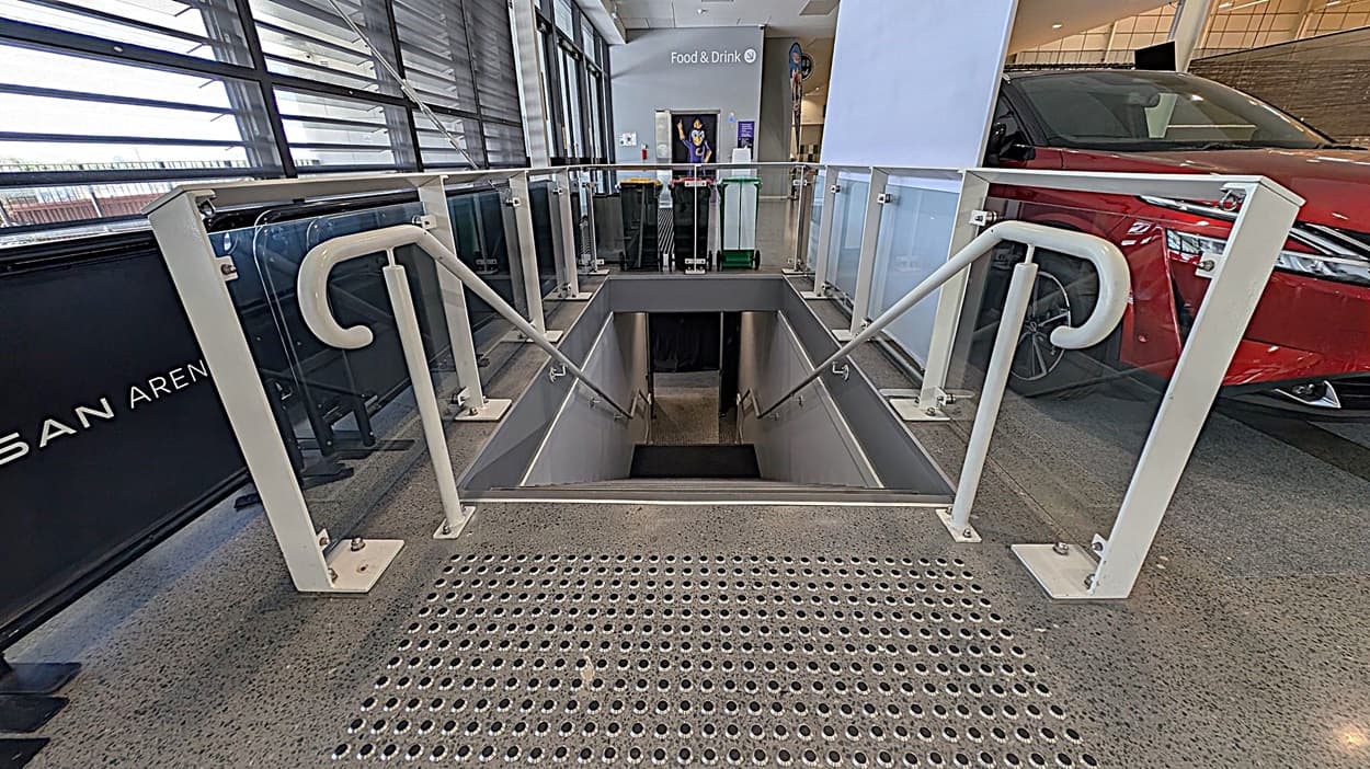 A staircase descends into a lower level, flanked by white railings and glass panels. The floor is speckled with a textured metal surface at the top of the stairs. To the right, a red car is partially visible. In the background, a sign reads "Food & Drink" with bins and a poster nearby. Large windows with horizontal blinds are on the left, allowing natural light into the space.