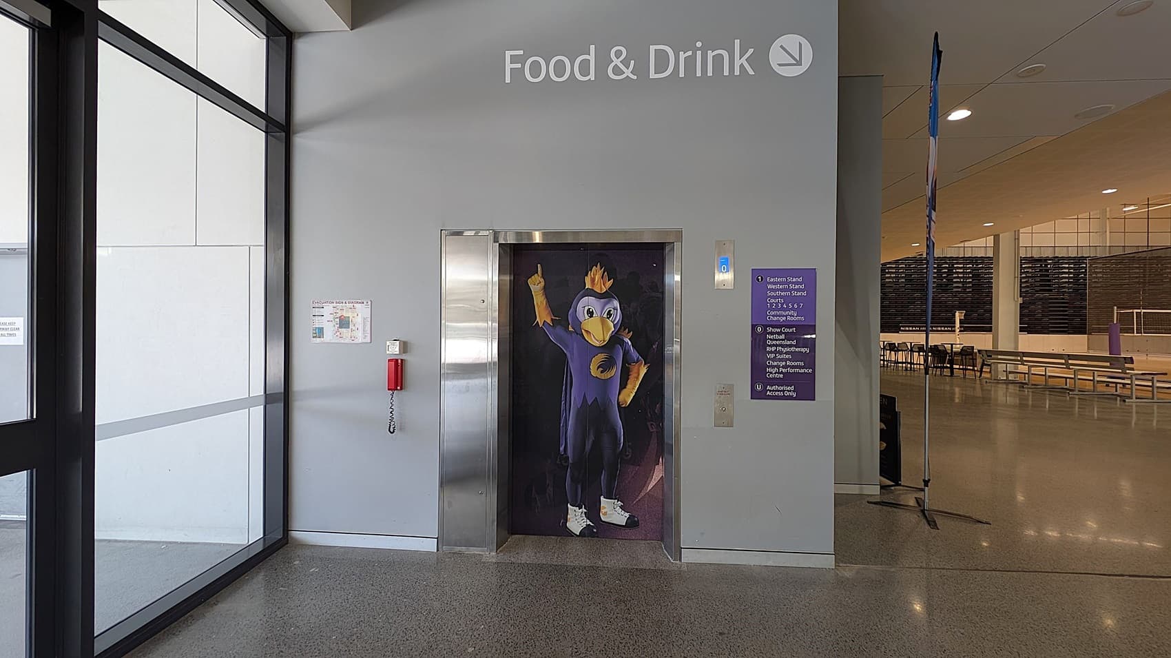 A hallway with a lift in the centre featuring a cartoon bird character on the door. Above, the words "Food & Drink" are displayed. To the left, large glass windows and a wall-mounted fire extinguisher. On the right, a sign with directions and a glimpse into a seating area with benches and tables. The floor is polished concrete, and the ceiling has recessed lighting.