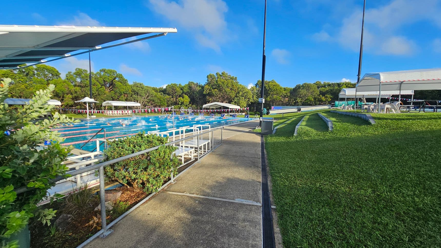 A swimming pool with multiple lanes is on the left, surrounded by greenery and shaded by a large canopy. Swimmers are visible in the water. On the right, there is a grassy area with tiered seating and a covered area with tables and chairs. The background features trees and a clear blue sky. A concrete path runs between the pool and the grassy area.
