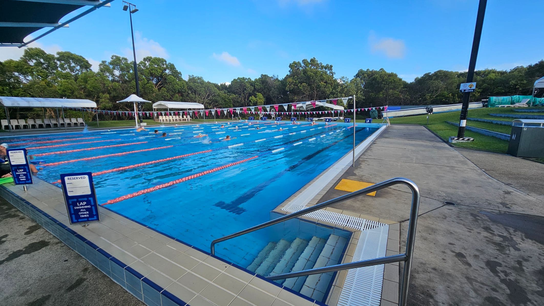 Outdoor swimming pool with multiple lanes, separated by orange lane dividers. Swimmers are in the water. On the left, there are signs indicating reserved lanes. The poolside is tiled, with steps leading into the water. In the background, there are white canopies and chairs, with trees beyond. On the right, a concrete path runs alongside the pool, with a grassy area and a tall light pole.