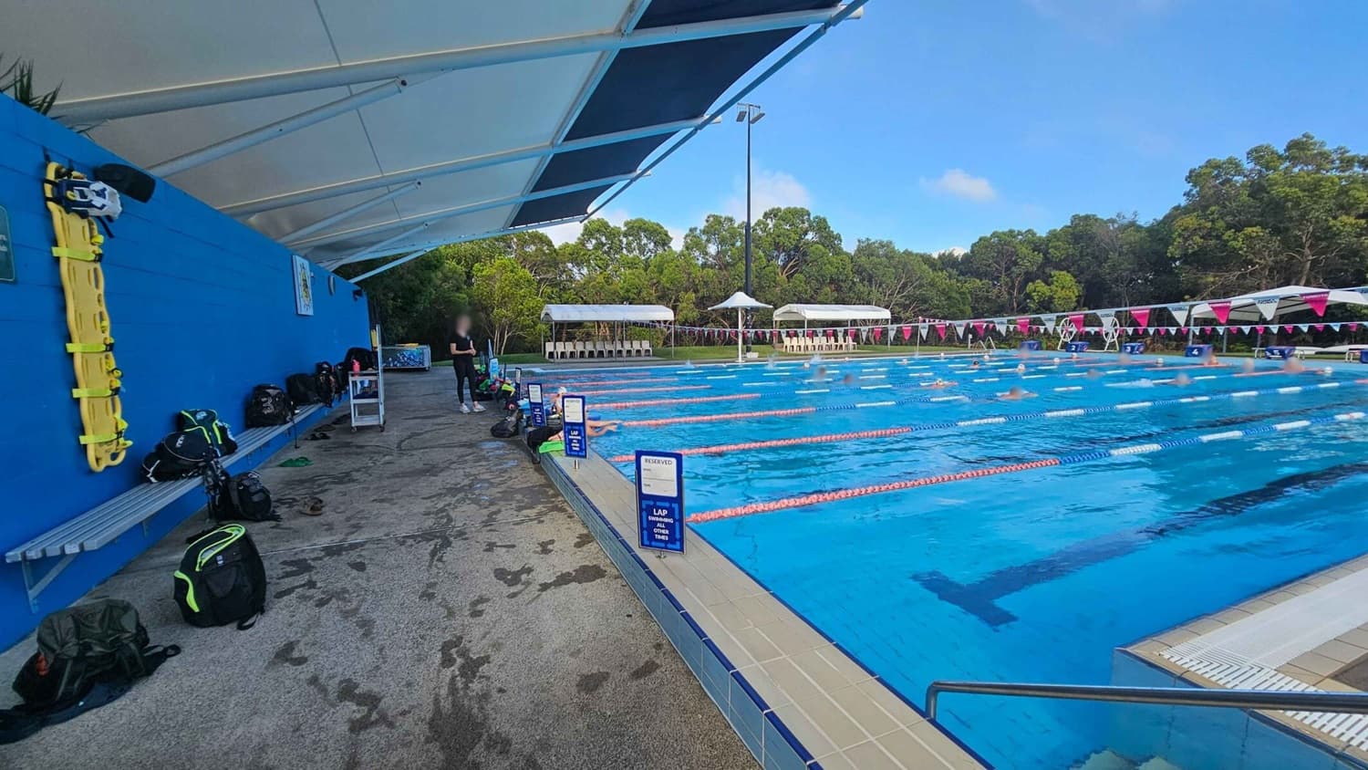 On the left, a blue wall with a yellow rescue board and several backpacks on a bench. The ground is wet. A white canopy covers the area. In the middle, a person stands near the edge of a swimming pool. On the right, a large outdoor pool with swimmers in designated lanes, marked by blue and red lane dividers. White signs indicate reserved lanes. In the background, trees and white seating areas with umbrellas are visible.
