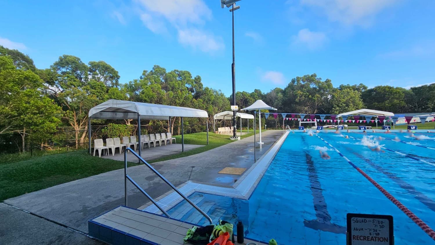 On the left, there are white plastic chairs under a canopy, with green trees in the background. In the middle, a concrete path runs alongside a swimming pool. The pool is on the right, with swimmers in lanes and colourful flags strung above. A sign with a schedule is visible in the foreground. The poolside area is wet, and there are some items like towels and bottles near the pool's edge.