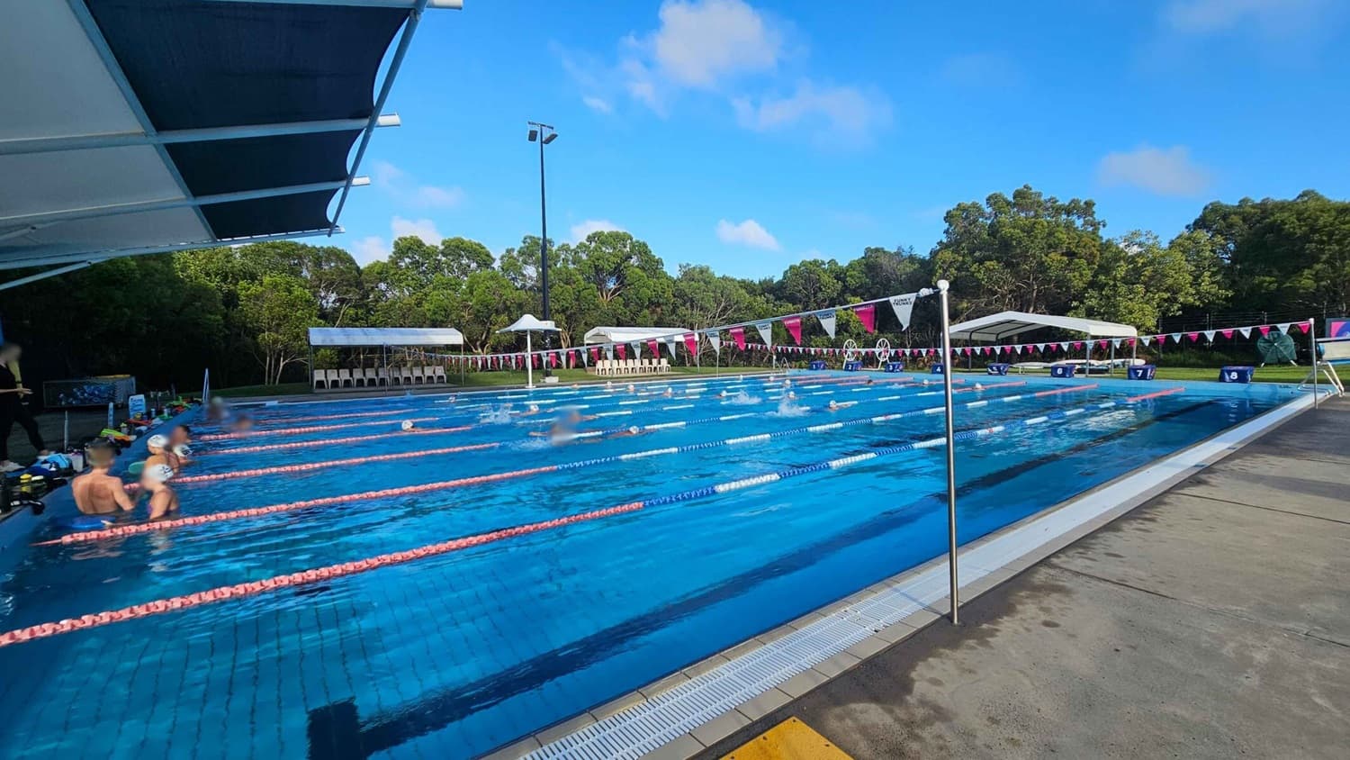 An outdoor swimming pool with multiple lanes, separated by pink lane dividers. Swimmers are in the water, with some resting at the edge on the left. A large canopy provides shade on the left side. In the background, there are white tents and chairs along the poolside, with trees and greenery beyond. The pool deck is concrete with a wet surface. Triangular flags are strung across the pool.