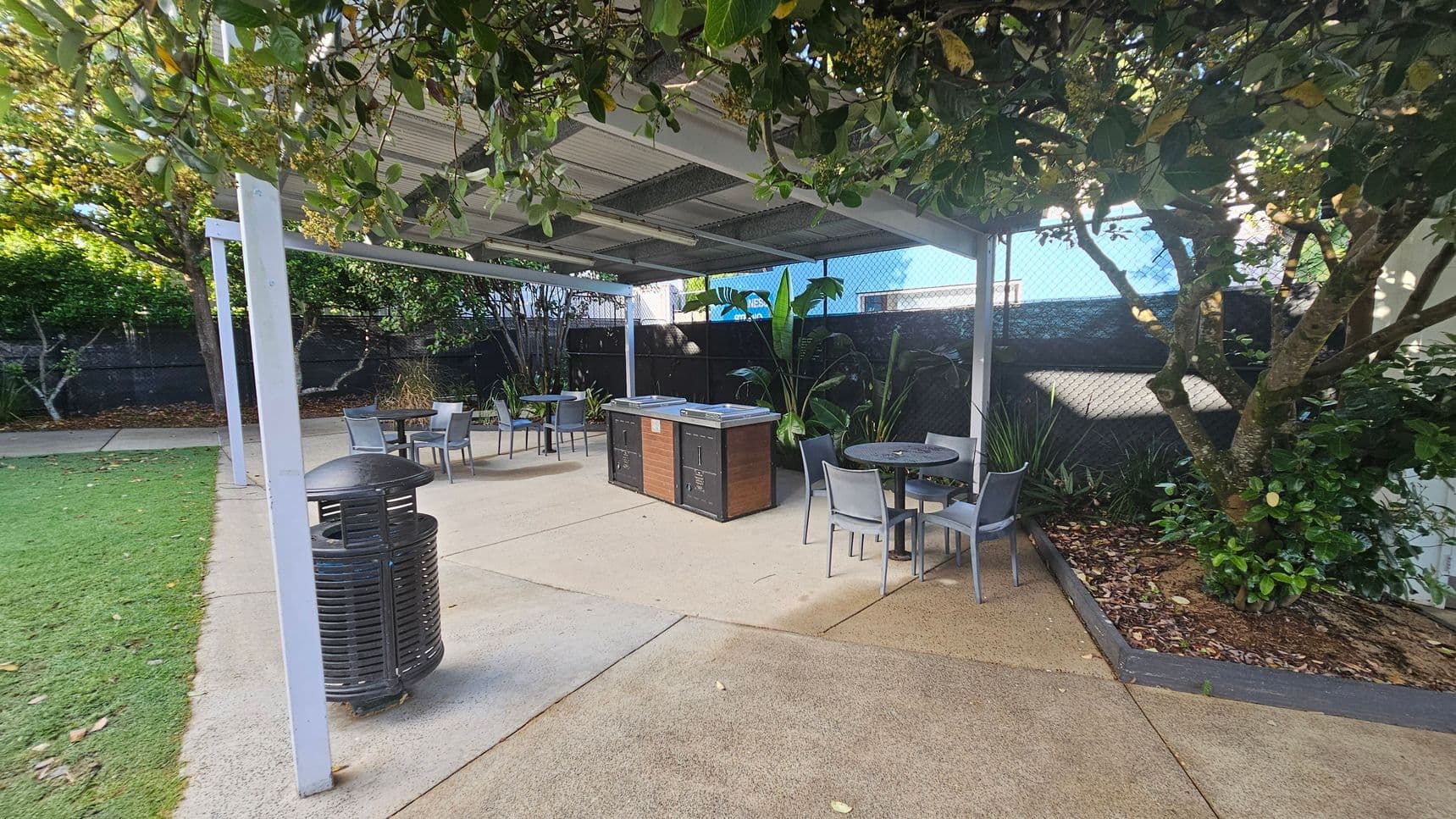 A covered outdoor area with a metal roof and white support beams. In the centre, there are two barbecue units and several round tables with grey chairs. The ground is a mix of concrete and grass. On the right, a tree with dense foliage provides shade, and there are various plants along the fence in the background. A black rubbish bin is on the left near the grass.