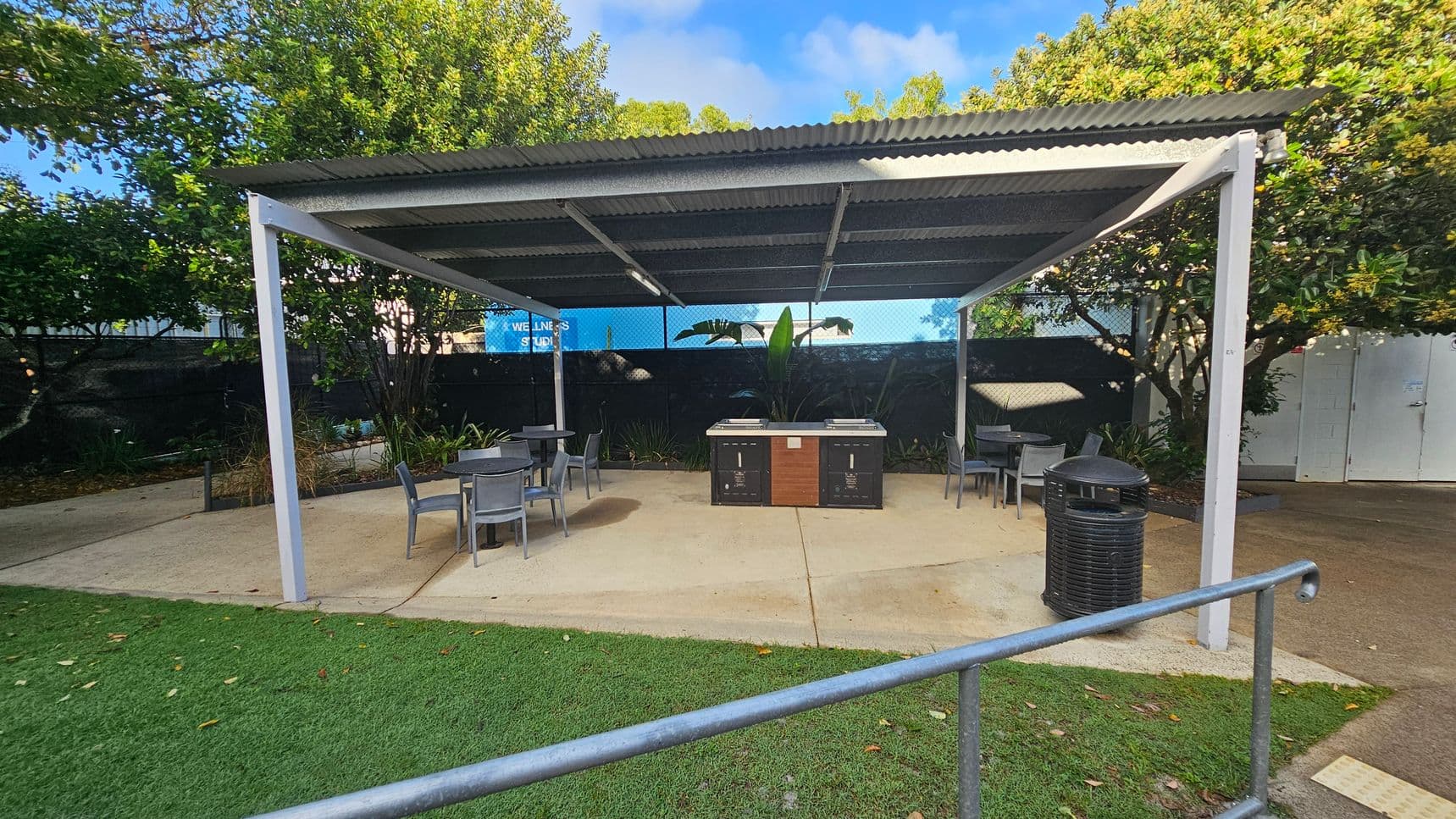 A covered outdoor area with a corrugated metal roof supported by white metal posts. In the centre, there are two barbecue grills with a wooden panel in between. Surrounding the grills are several grey tables and chairs. A black rubbish bin is on the right. The ground is a mix of concrete and artificial grass. In the background, there are trees and a blue sign partially visible, reading "WELLNESS STUDIO".