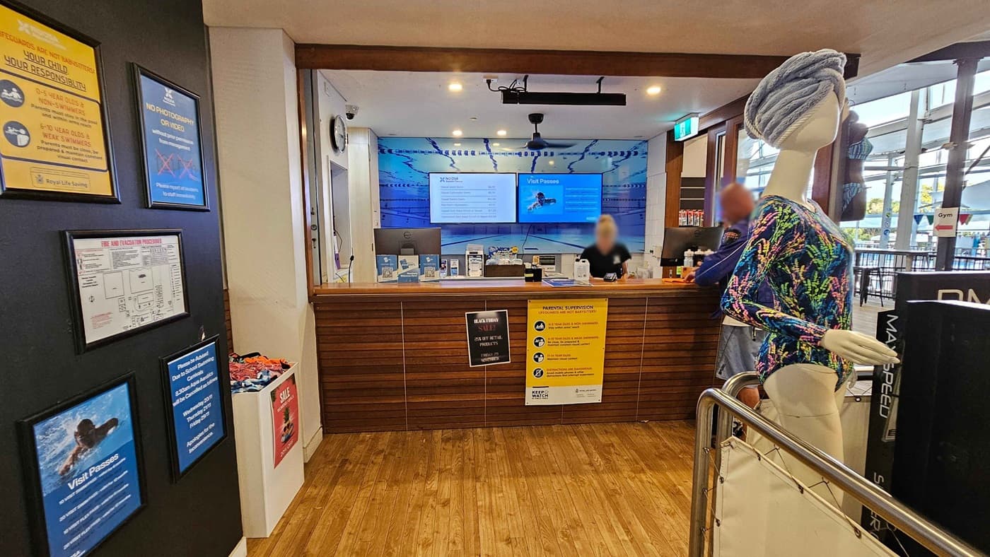 A café with a wooden interior. On the right, a barista is preparing drinks behind a counter with a display of pastries and a coffee machine. Shelves hold coffee supplies and a sign for coconut water. In the centre, a TV screen is mounted above the counter. On the left, an ice cream freezer with "Peters" branding is next to a display of clothing and accessories. The floor is wooden, and the ceiling is white with recessed lighting.