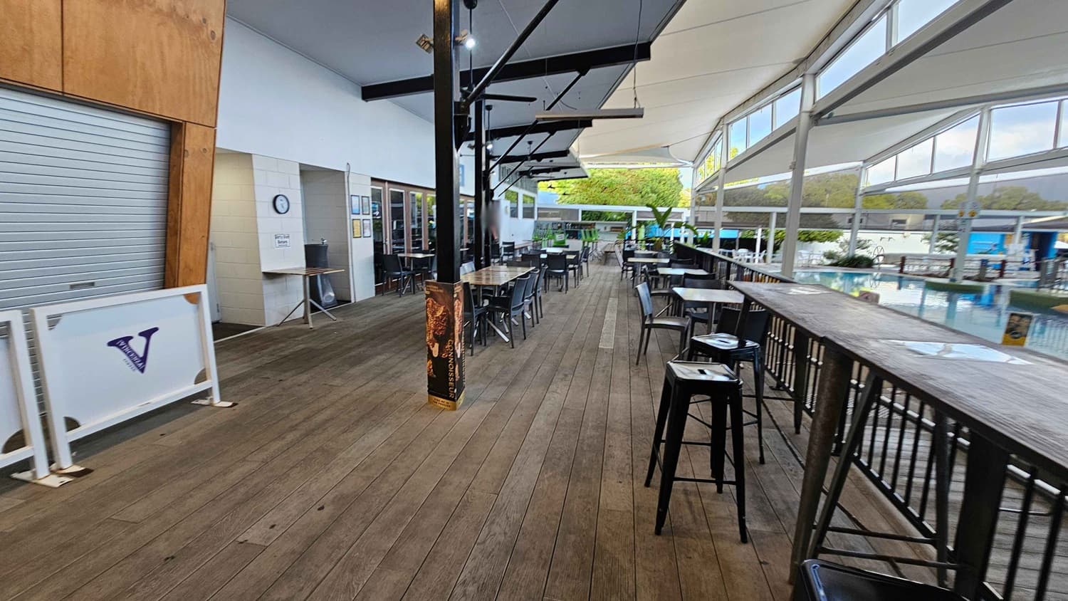 A spacious outdoor seating area with wooden flooring. On the left, there's a closed metal shutter and a sign with a "V" logo. In the middle, several tables and chairs are arranged under a canopy with black support beams. On the right, a long wooden bar with high stools overlooks a pool area. Large windows and a white ceiling allow natural light to fill the space. Greenery is visible in the background near the pool.