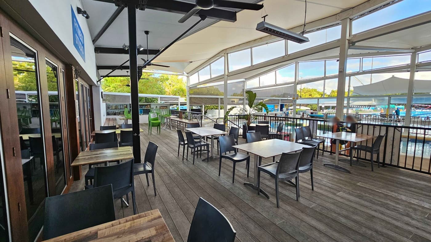 Outdoor seating area with wooden tables and black chairs on a wooden deck. On the left, glass doors reflect the surroundings. In the middle, tables are arranged in rows, with some stacked green chairs in the background. The right side features a railing overlooking a pool area with people visible in the distance. The ceiling is covered with a white canopy, and large windows allow natural light to fill the space.