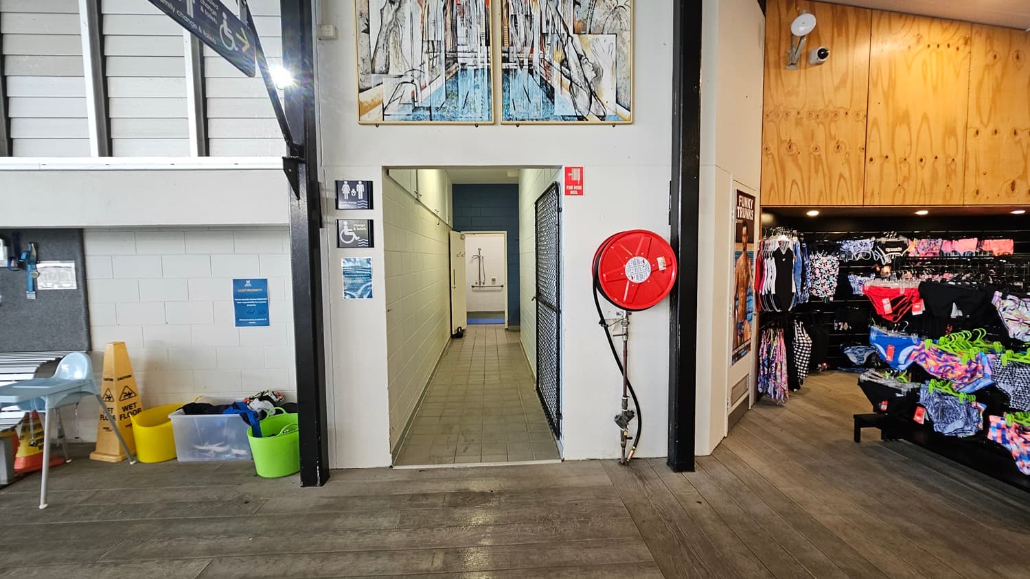 A corridor with tiled flooring leads to a changing area. On the left, there are chairs, wet floor signs, and storage bins. On the right, a red fire hose reel is mounted on the wall, and a shop area displays swimwear on racks. The walls are white with signage for accessible toilets. Above the corridor entrance, there is a colourful artwork. The ceiling is wooden on the right side.