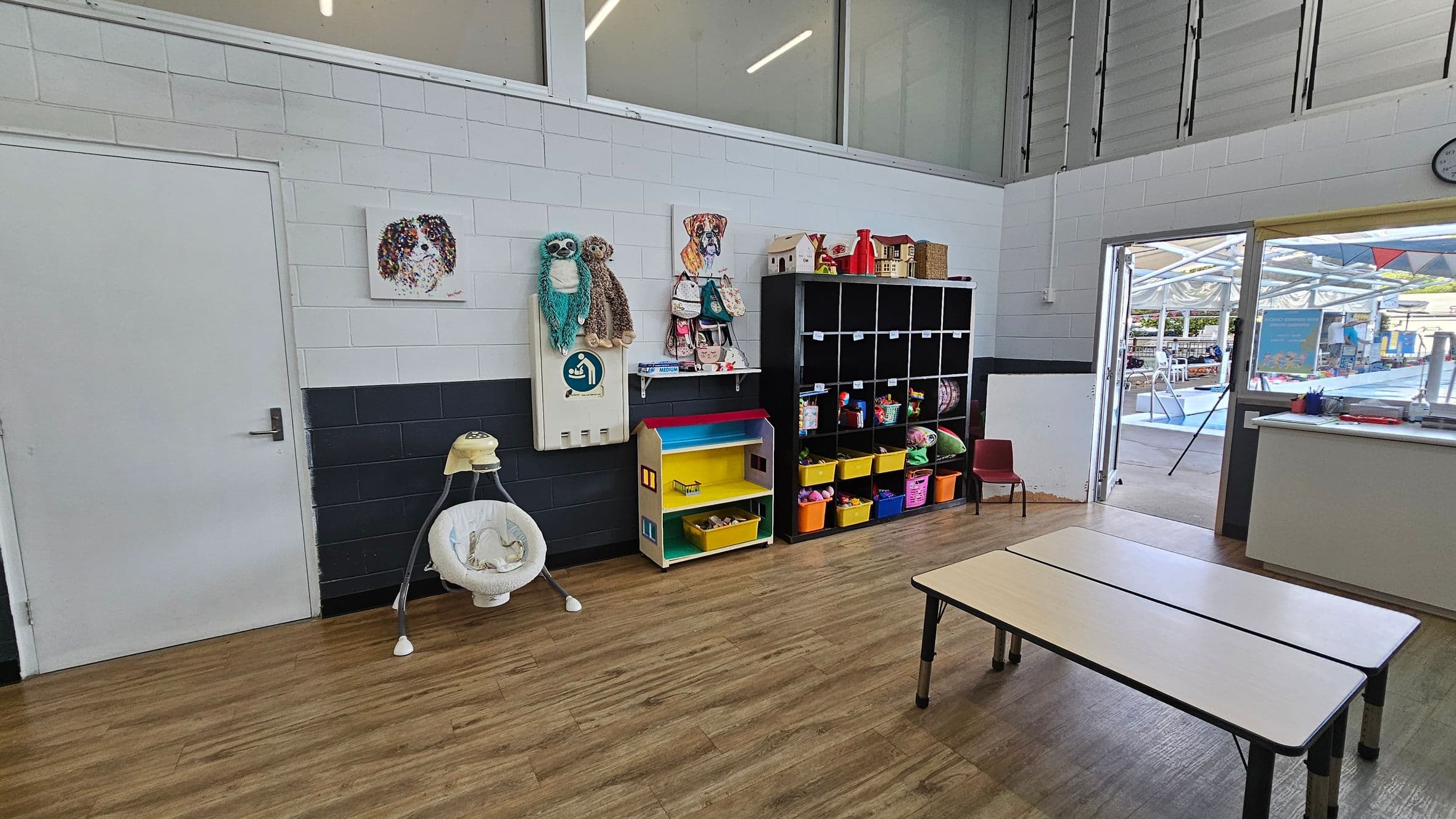 A classroom with a wooden floor and white ceiling. On the left, there's a white door and a baby swing. The wall has a painting of a dog and two plush toys hanging. In the middle, a colourful shelf with yellow bins and a black cubby unit filled with toys. On the right, two white tables and a small red chair. A door leads outside to a covered area with a pool. A clock is visible on the wall near the door.