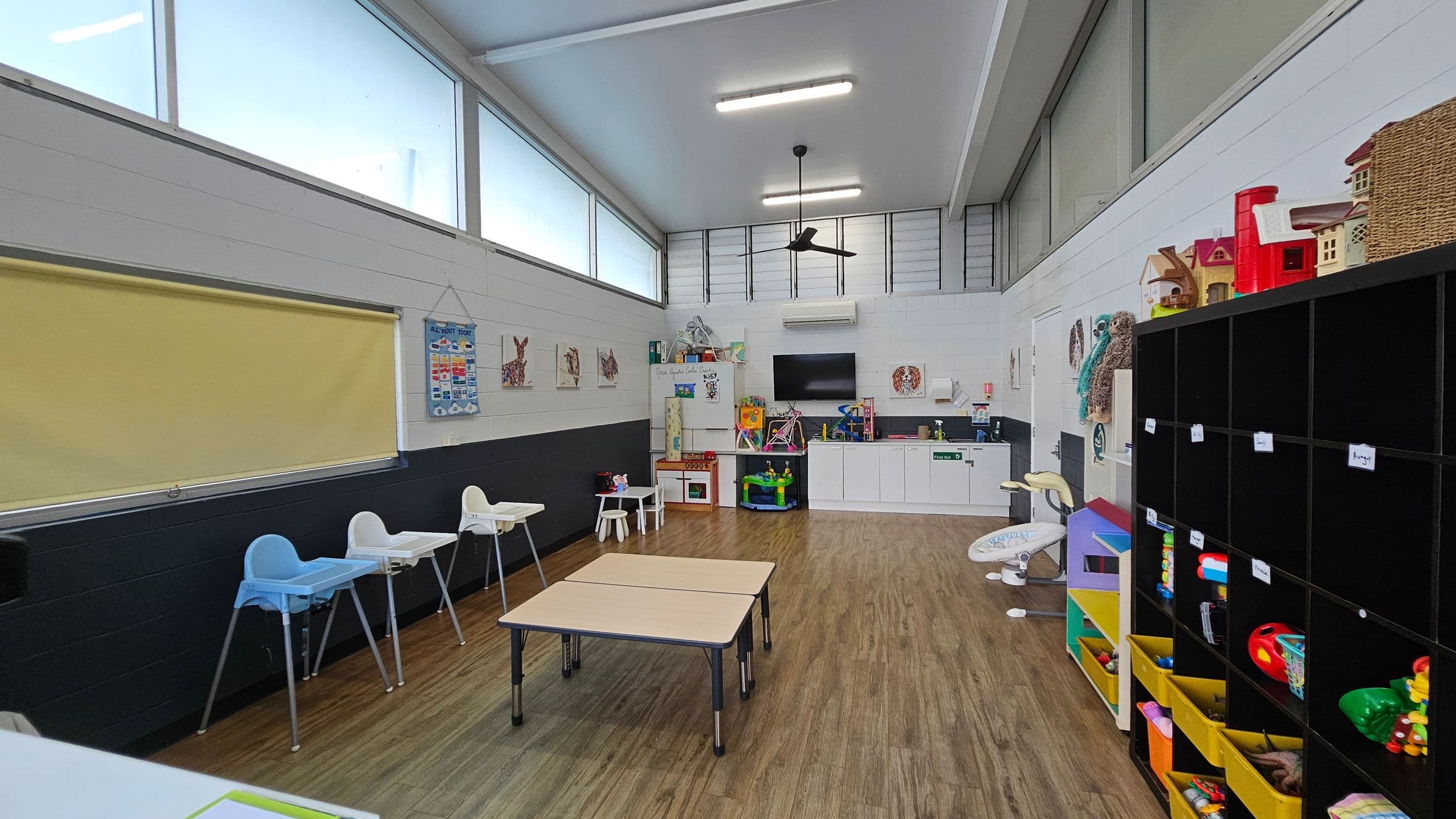 A brightly lit playroom with large windows near the ceiling. On the left, there are high chairs and a yellow blind. In the centre, two small tables are placed on a wooden floor. The back wall features a kitchenette with a TV above it, and various toys and educational materials. On the right, a black cubby shelf holds toys and baskets. The ceiling has fluorescent lights and a ceiling fan.