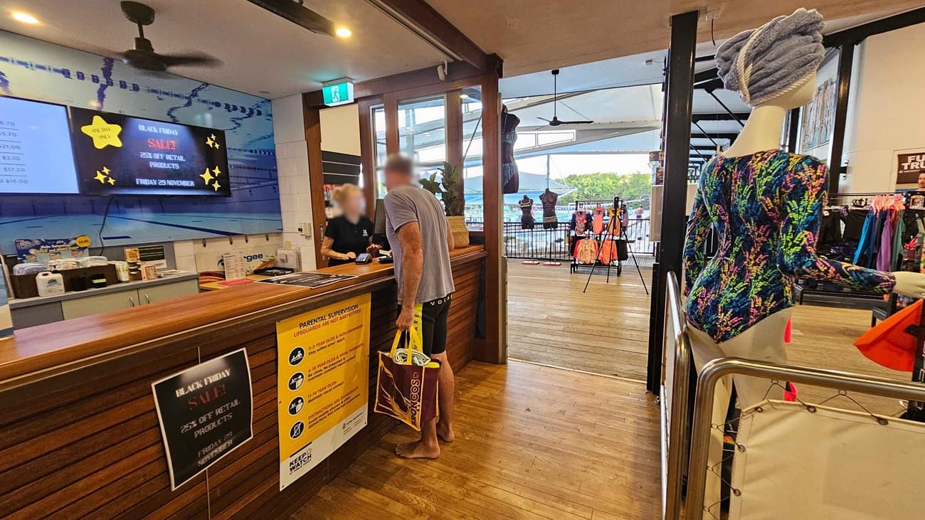 To the left, a wooden counter features a sales sign and various pricing boards above it, with a staff member assisting a customer. On the right, a mannequin dressed in a colourful swimsuit stands beside a display of swimwear and accessories. In the background, large windows allow light to filter through, revealing more swimwear on racks. The floor has a polished wooden surface, and the ceiling is plain with several light fixtures and ceiling fans visible