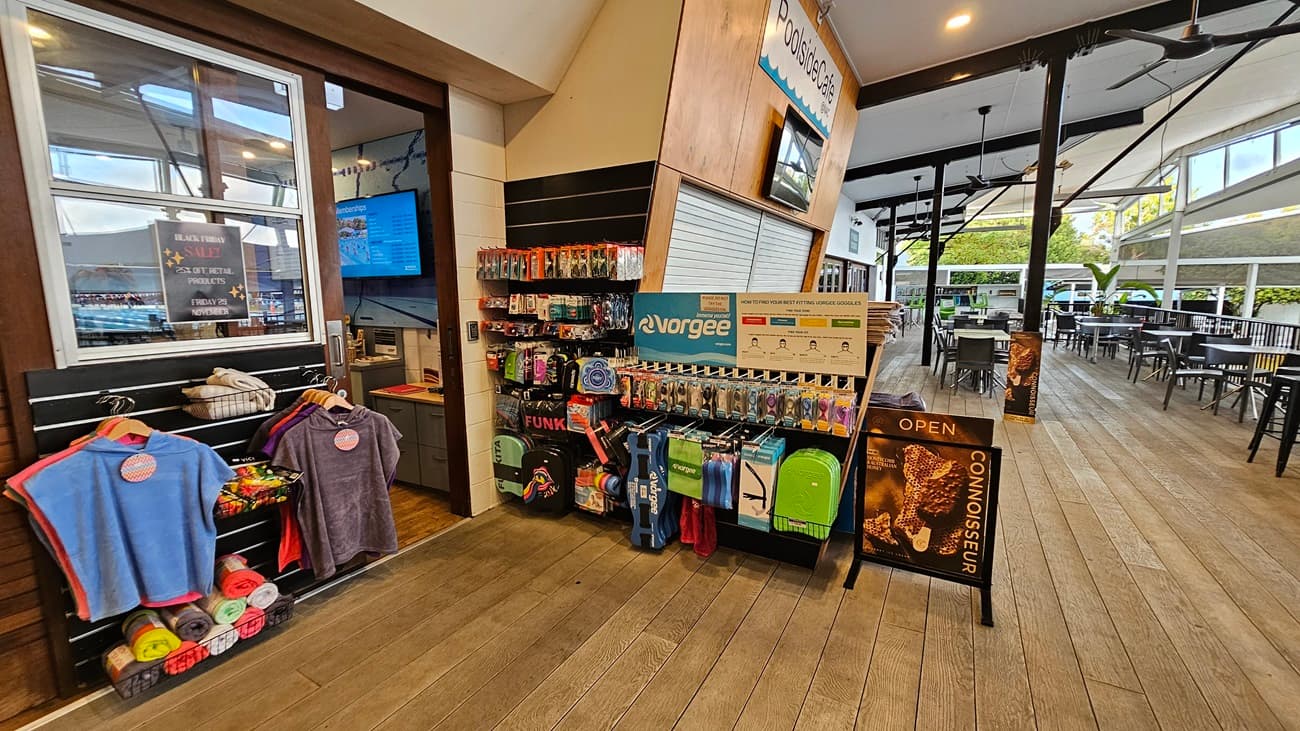 To the left, a display features a variety of colourful towels hung on racks, with a sign indicating the café nearby. Next to it, a wall-mounted screen shows information, and the counter displays various products for sale. On the right, a range of swimming accessories and bags is neatly organised on shelves, with an “Open” sign visible. The wooden floor runs throughout the area, while the ceiling has exposed beams and ceiling fans, contributing to an open and airy atmosphere.